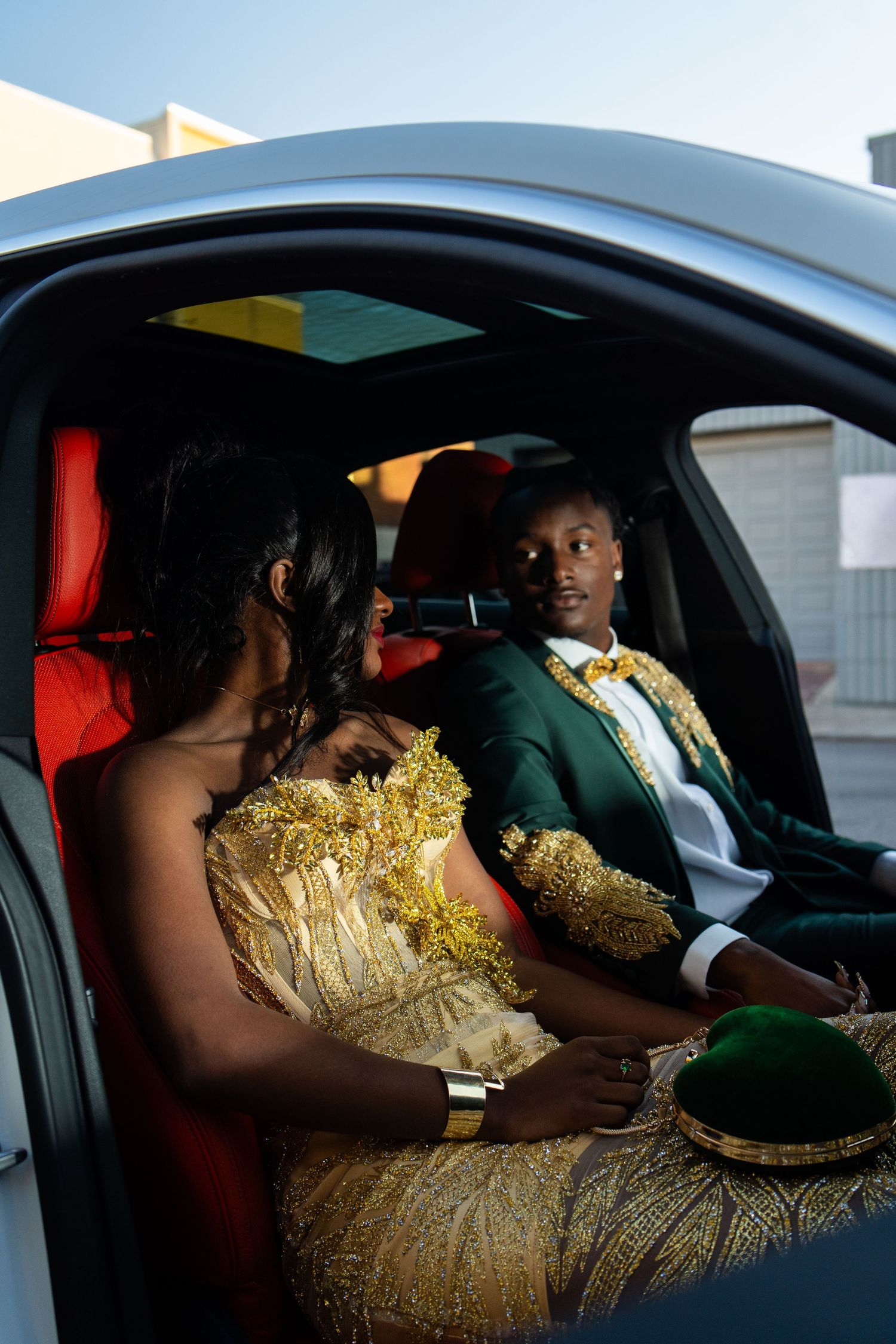 A couple sits in formal attire inside a luxury car with gold accents and red interior.