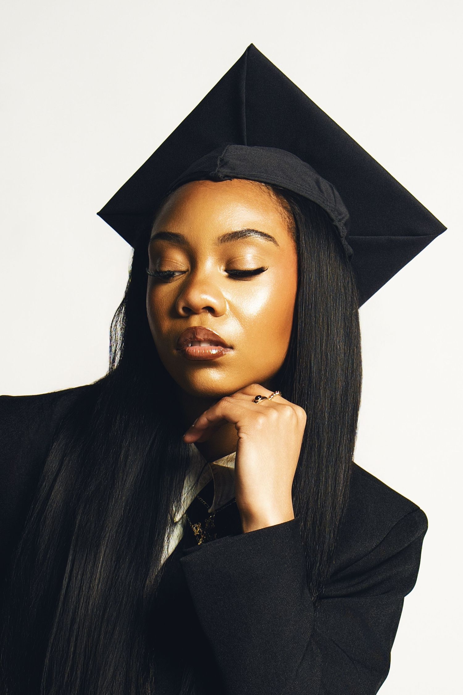 Young Black woman in graduation cap and gown poses thoughtfully with eyes closed against white background.