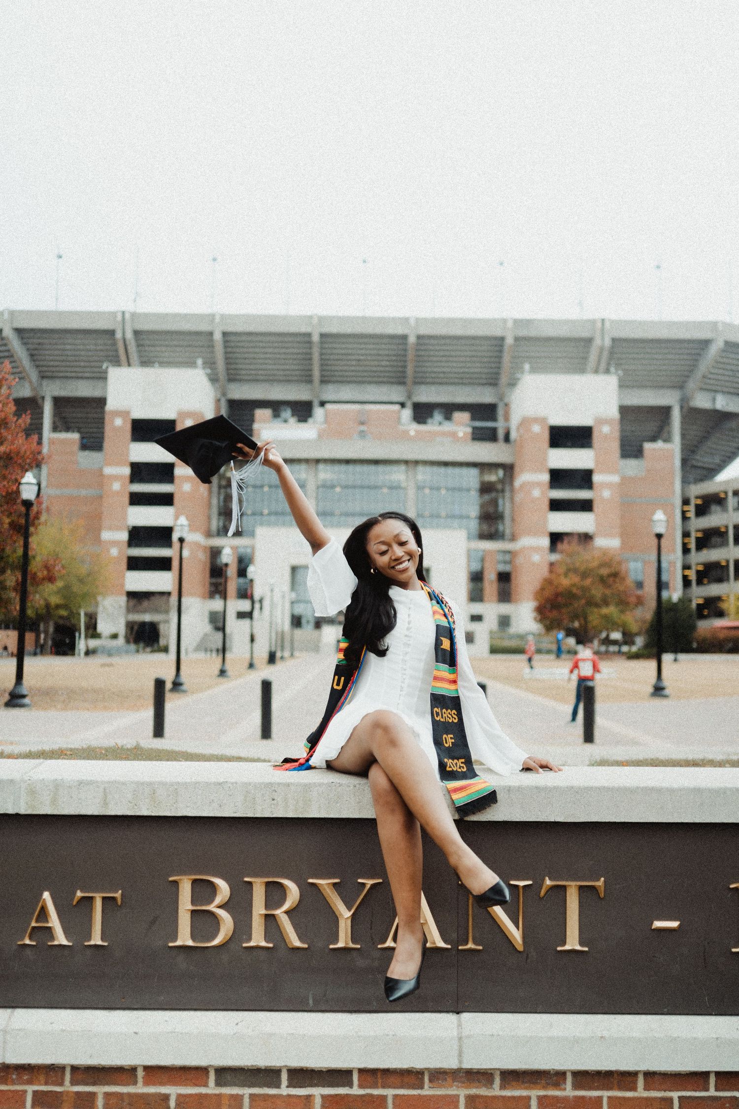 Graduate in white dress sitting on Bryant sign outside stadium during autumn graduation photoshoot.