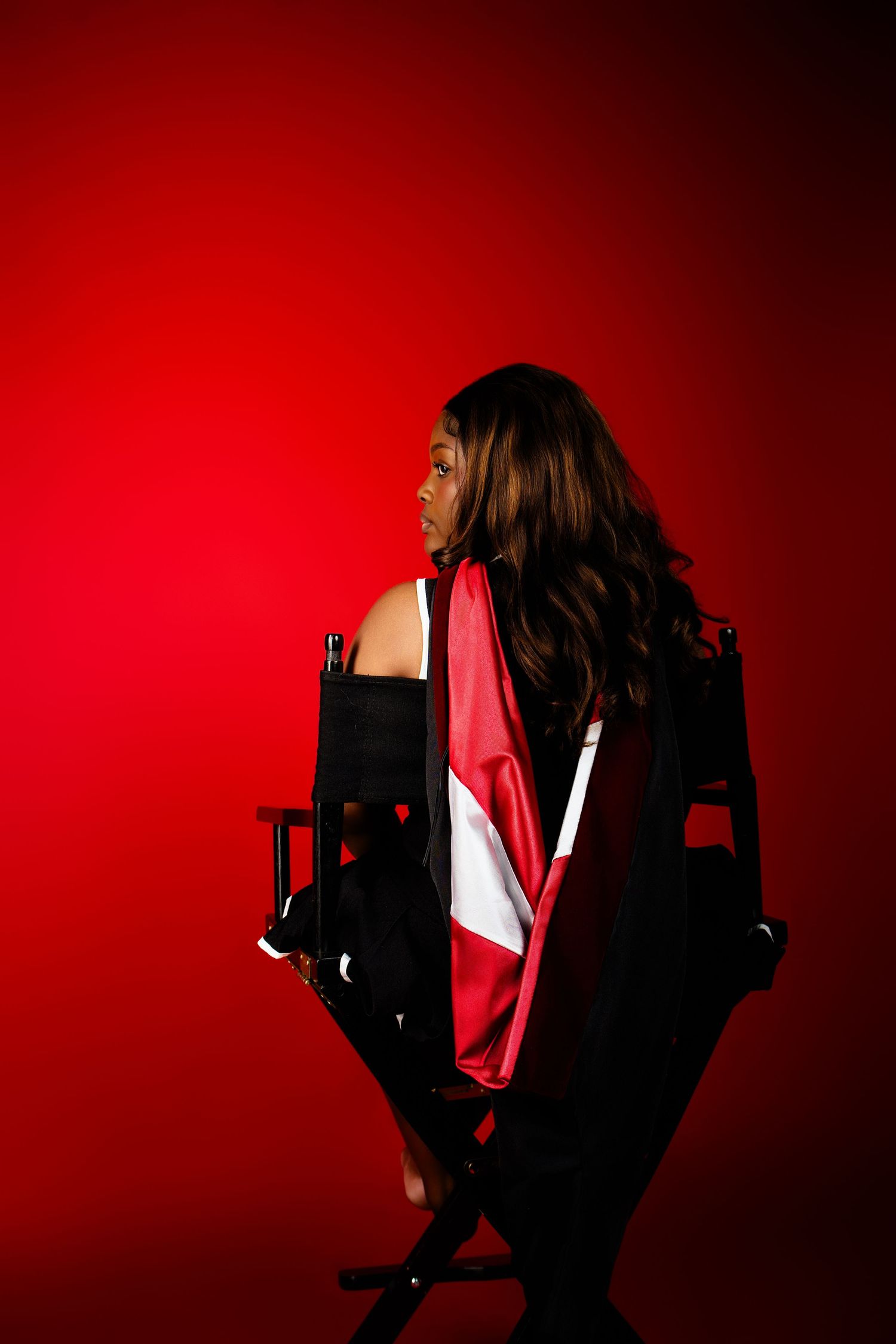 Woman with long dark hair draped in a red and white flag sits in a director's chair against a bold red background.