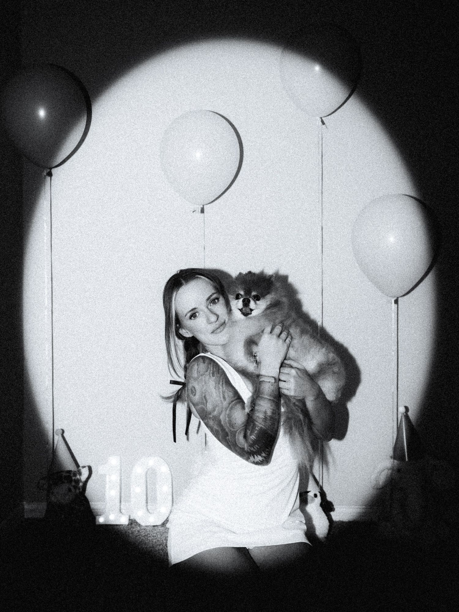 Woman with tattoos holding a Pomeranian dog, surrounded by balloons and a number 10, in black and white photo.