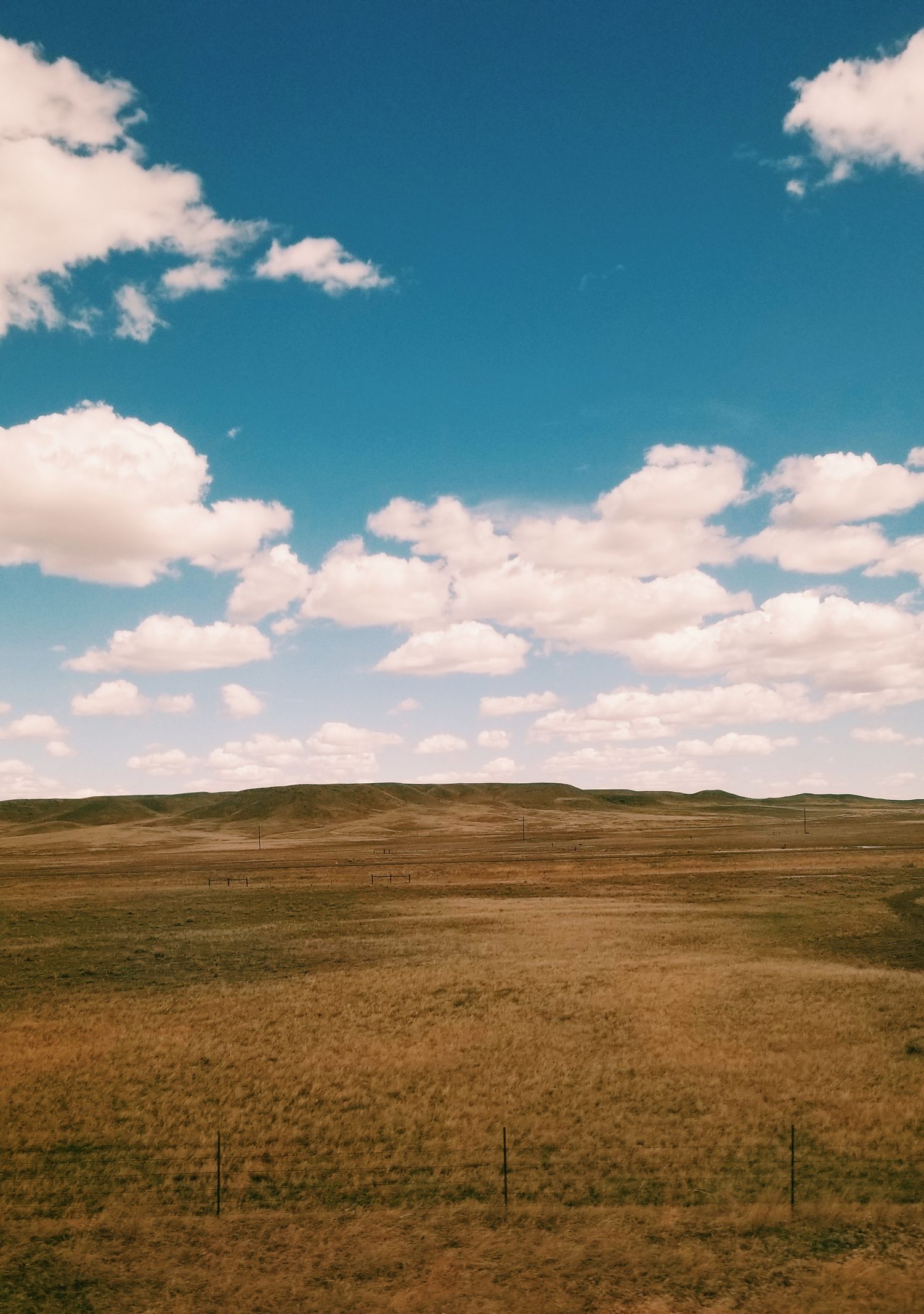 A panoramic view of rolling brown prairie grasslands under a bright blue sky with scattered white clouds.