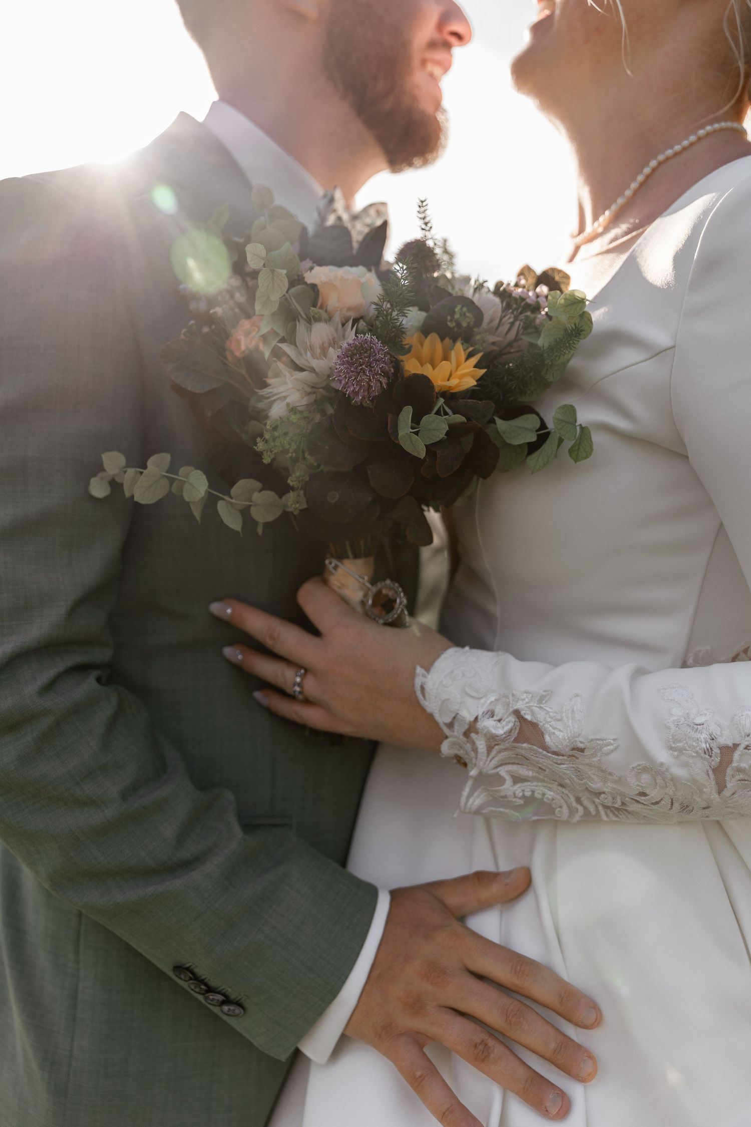 Bride and groom embracing outdoors, holding a rustic wildflower bouquet with sunflowers and eucalyptus in golden sunlight.