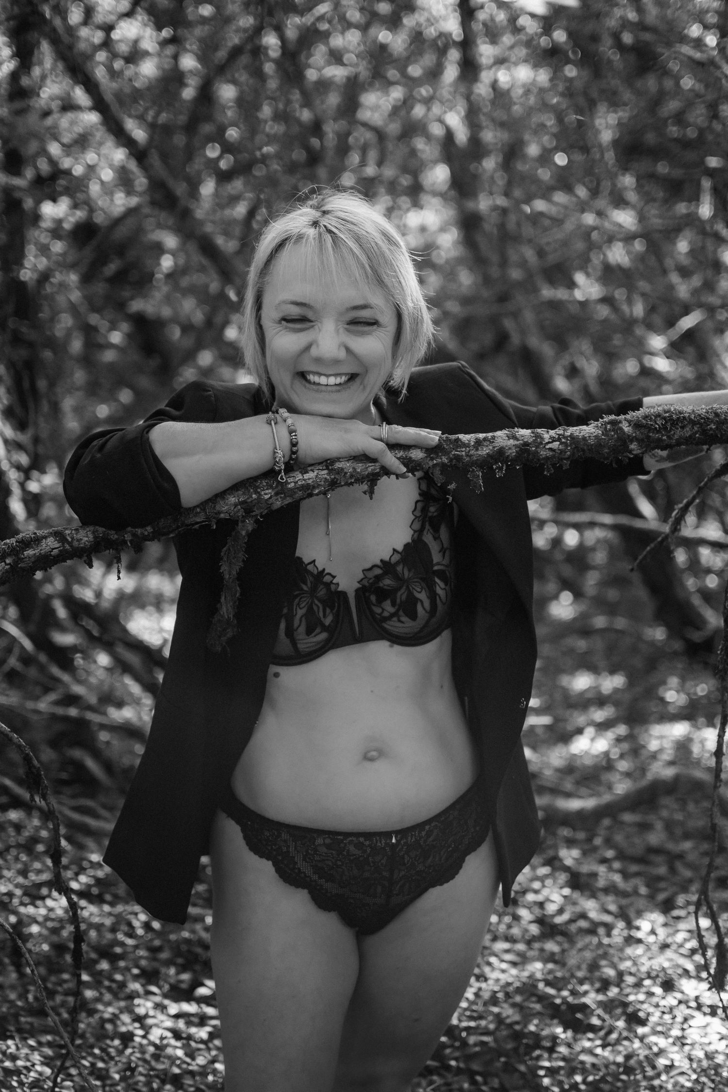 Smiling blonde woman in lace lingerie and blazer leaning on a tree branch in a forest, black and white photo.