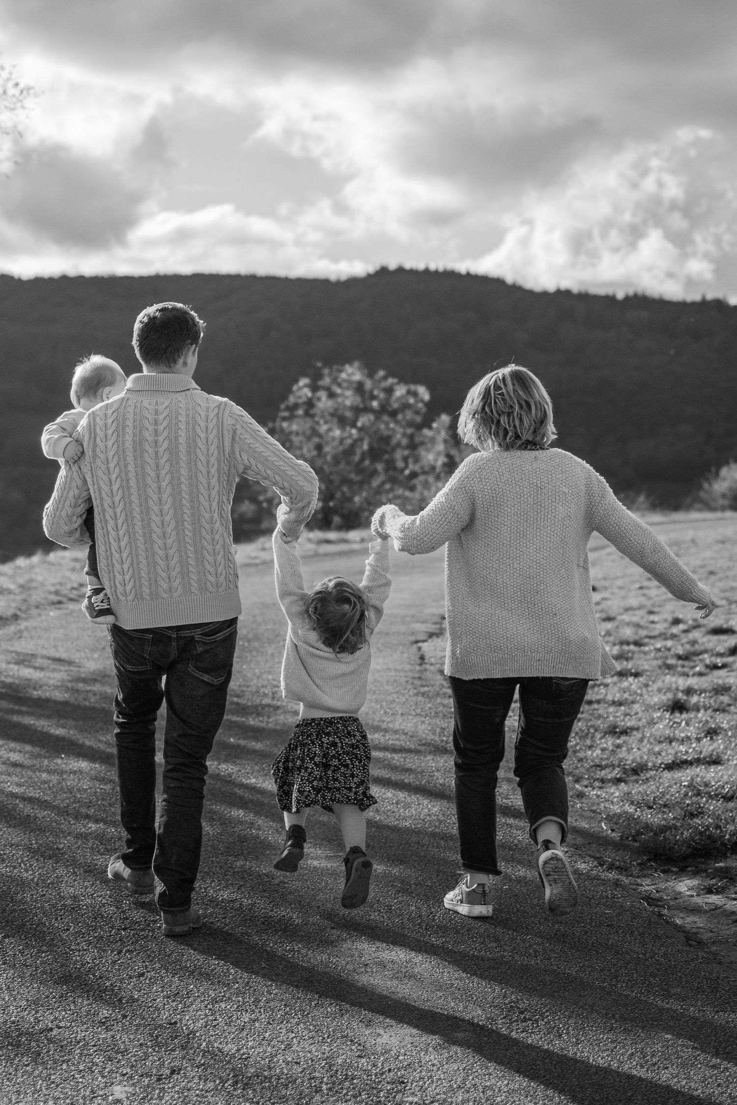 Black and white photo of a family walking down a rural road, parents holding children's hands with hills in background.