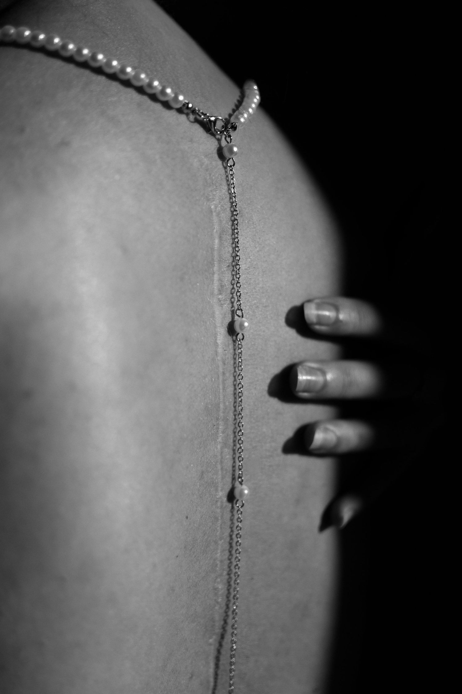 Black and white close-up of a womans bare back adorned with a pearl and chain necklace, with a hand resting on her shoulder.