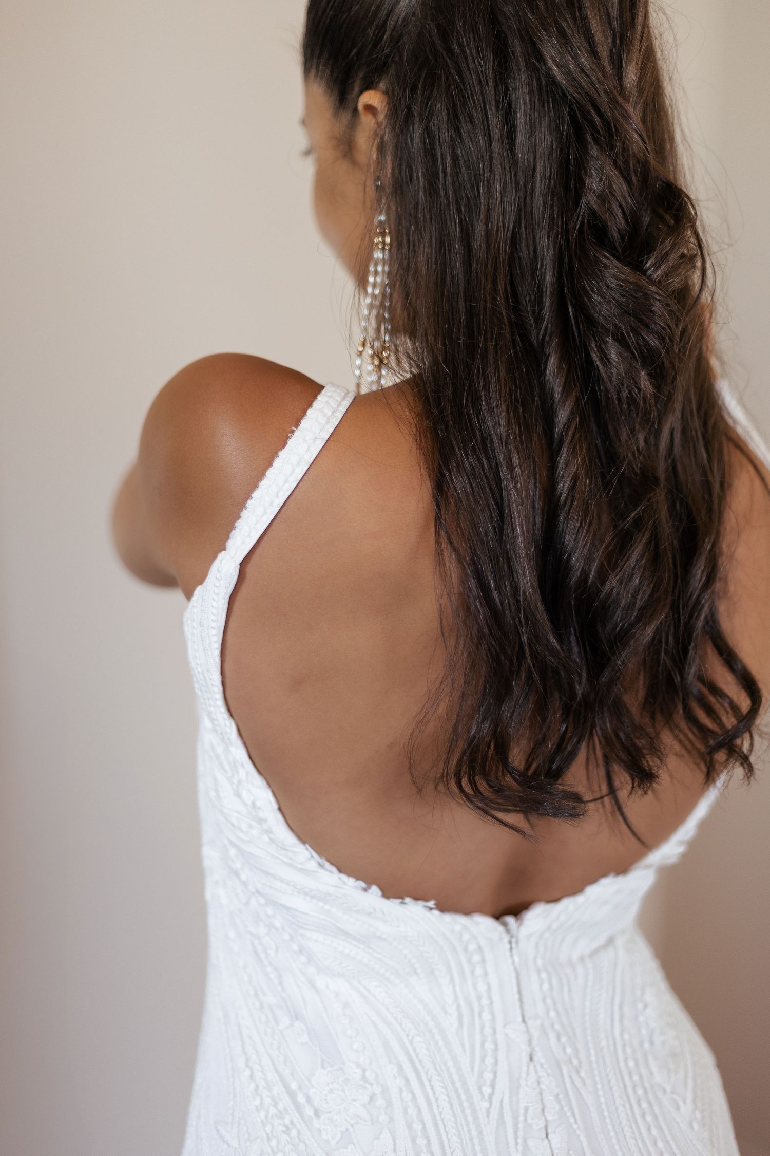 Woman in white beaded backless dress with long dark hair and crystal drop earrings, elegant bridal fashion.