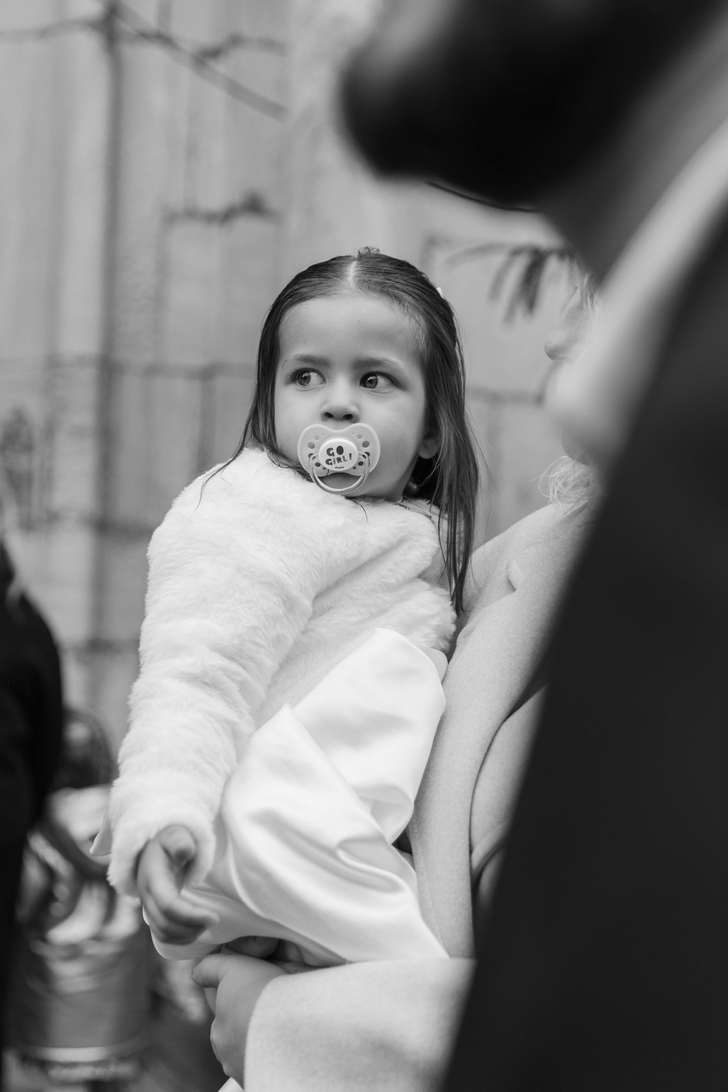Black and white photo of a young girl with a pacifier, wrapped in a white fluffy robe, being held by an adult.