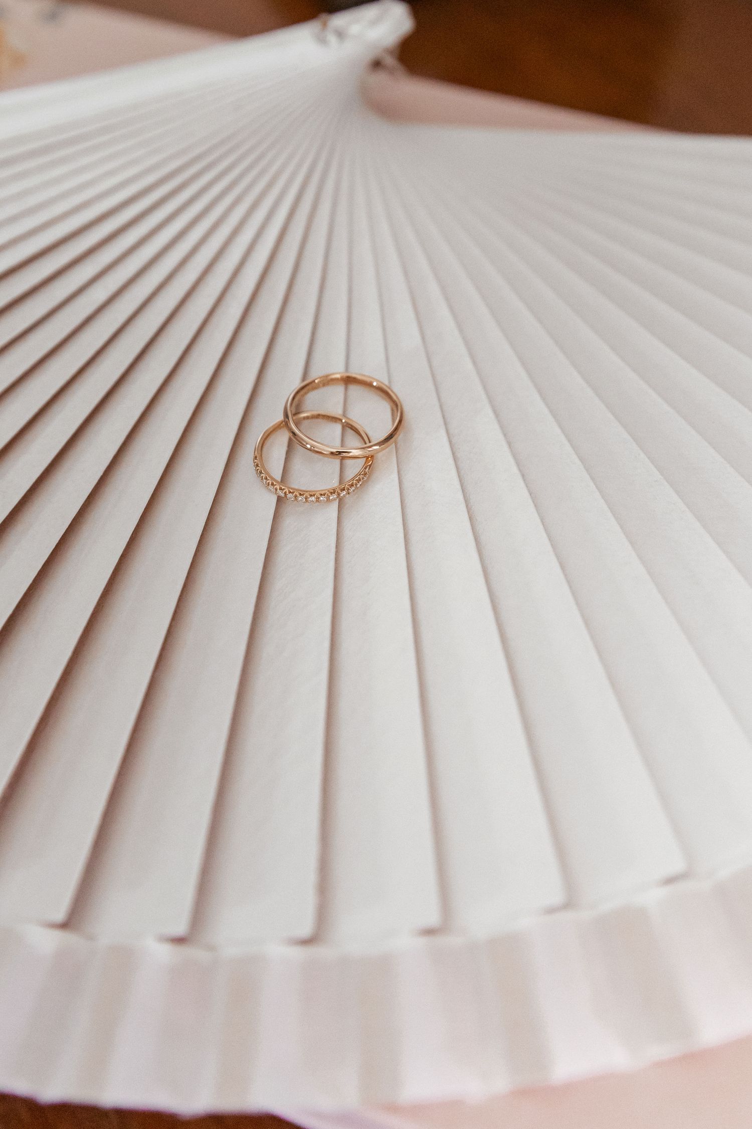 Two gold wedding bands resting on a white pleated paper fan, elegant bridal accessories detail shot.