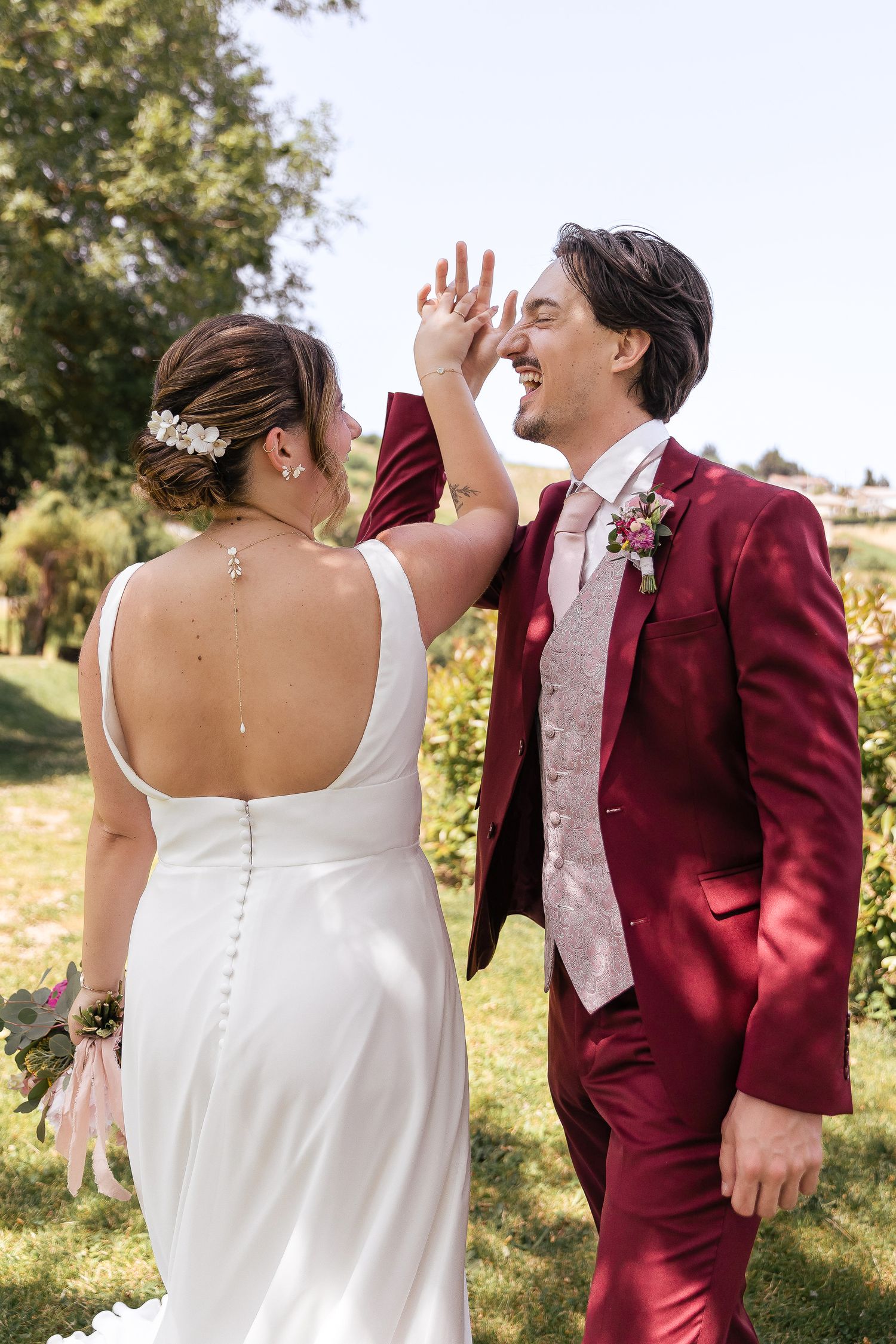 A wedding couple in burgundy suit and white dress share candid laughing moments during an outdoor photo session.