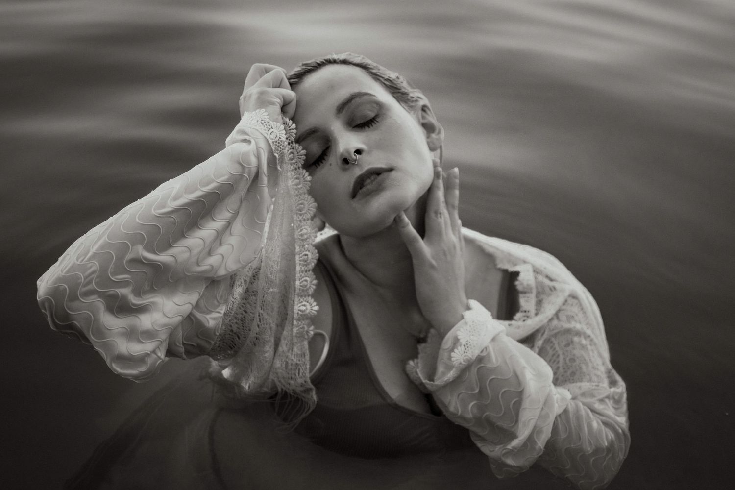 Black and white photo of a woman in lace emerging from water, hands gracefully touching her face.