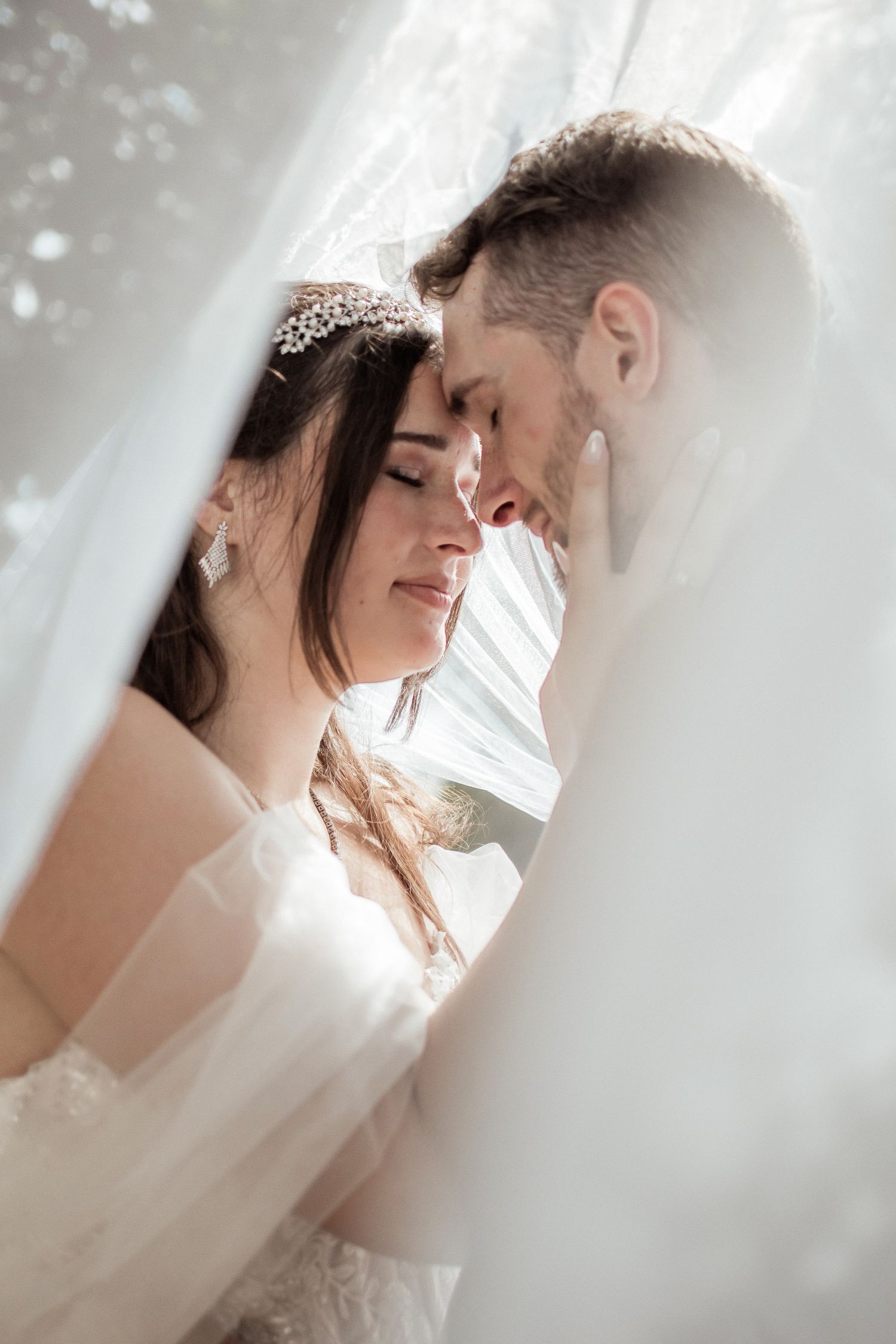 A romantic wedding couple shares an intimate moment behind a soft white veil in a series of ethereal photos.