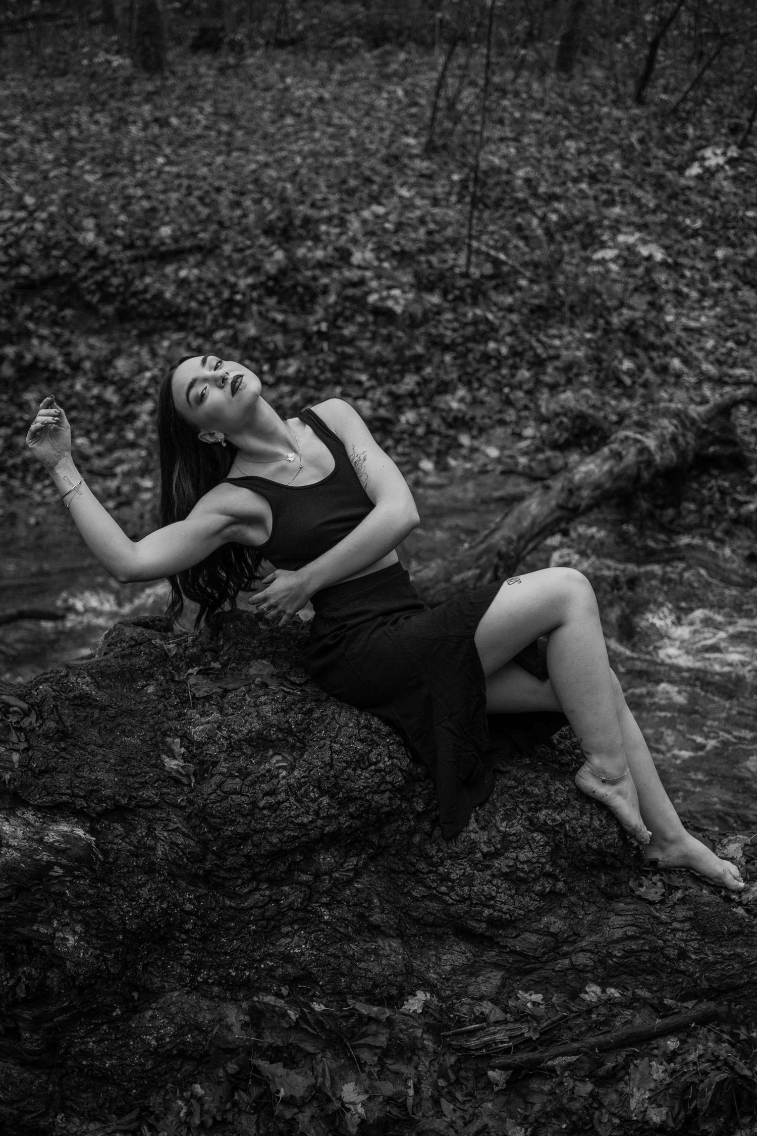 Black and white photo of a woman in a dark dress reclining gracefully on mossy rocks beside a forest stream.