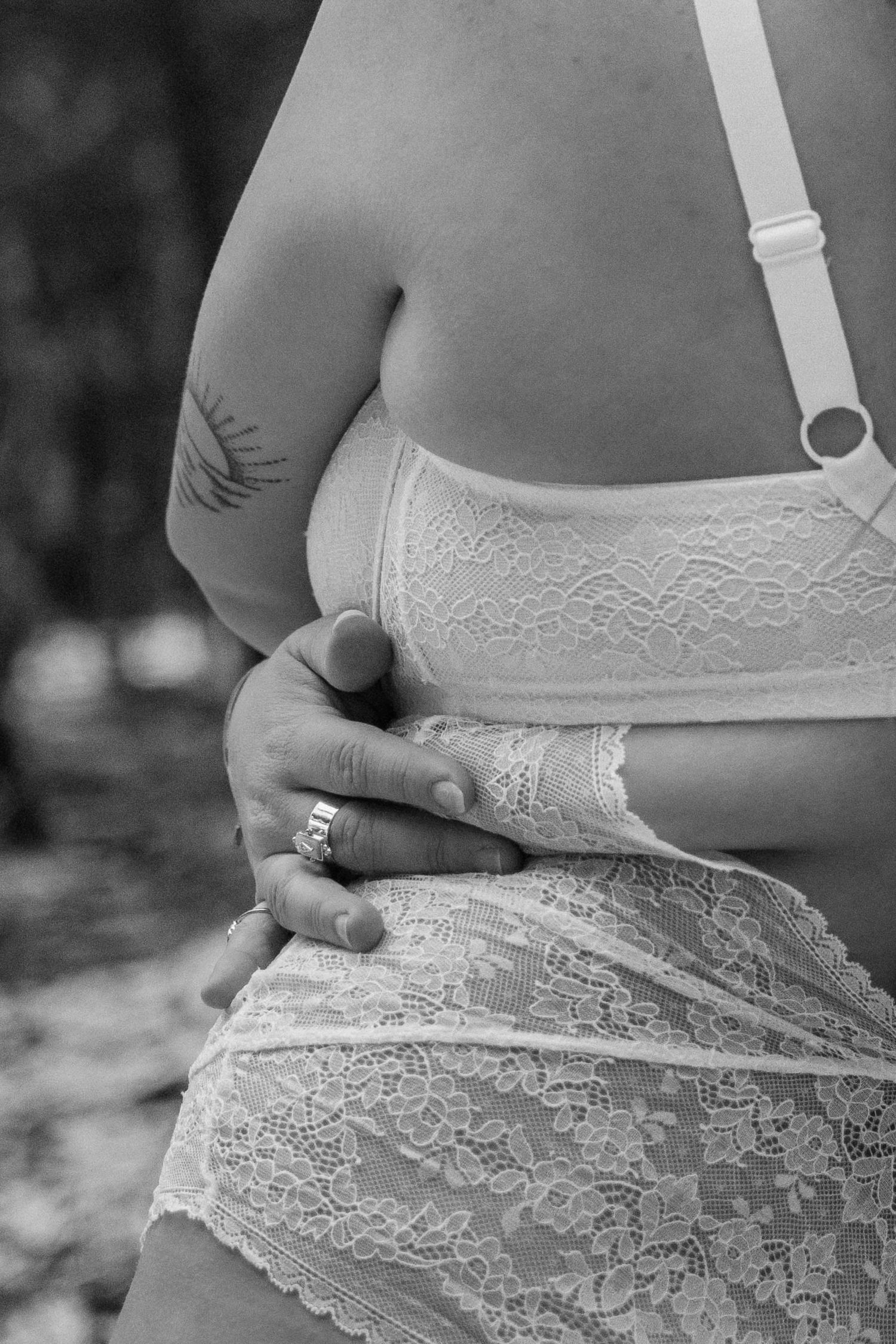 Black and white photo of a woman in lace lingerie with a tattoo, hands clasped at her waist.