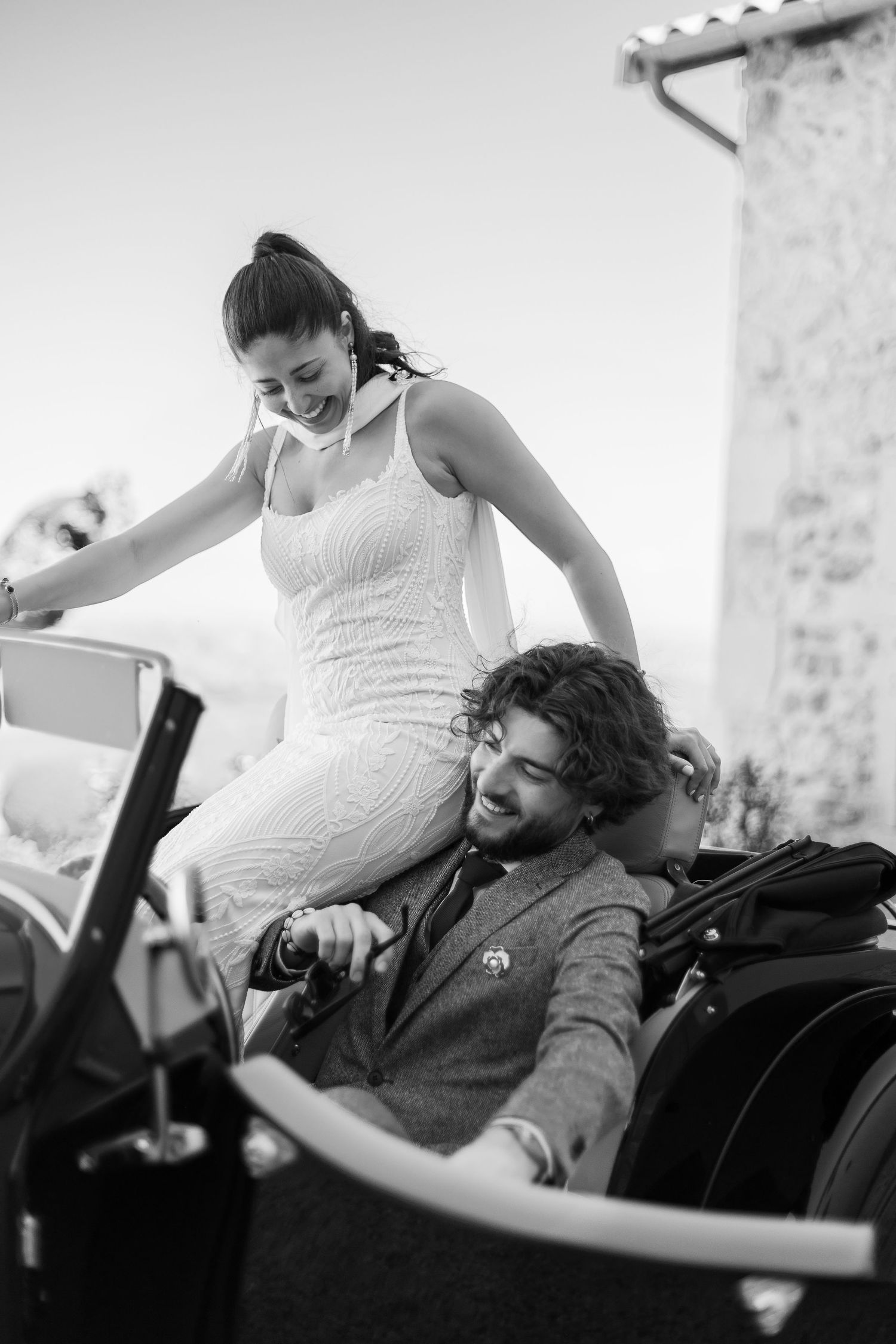 Black and white photo of a smiling bride in white dress and groom in suit sitting in a vintage convertible car.