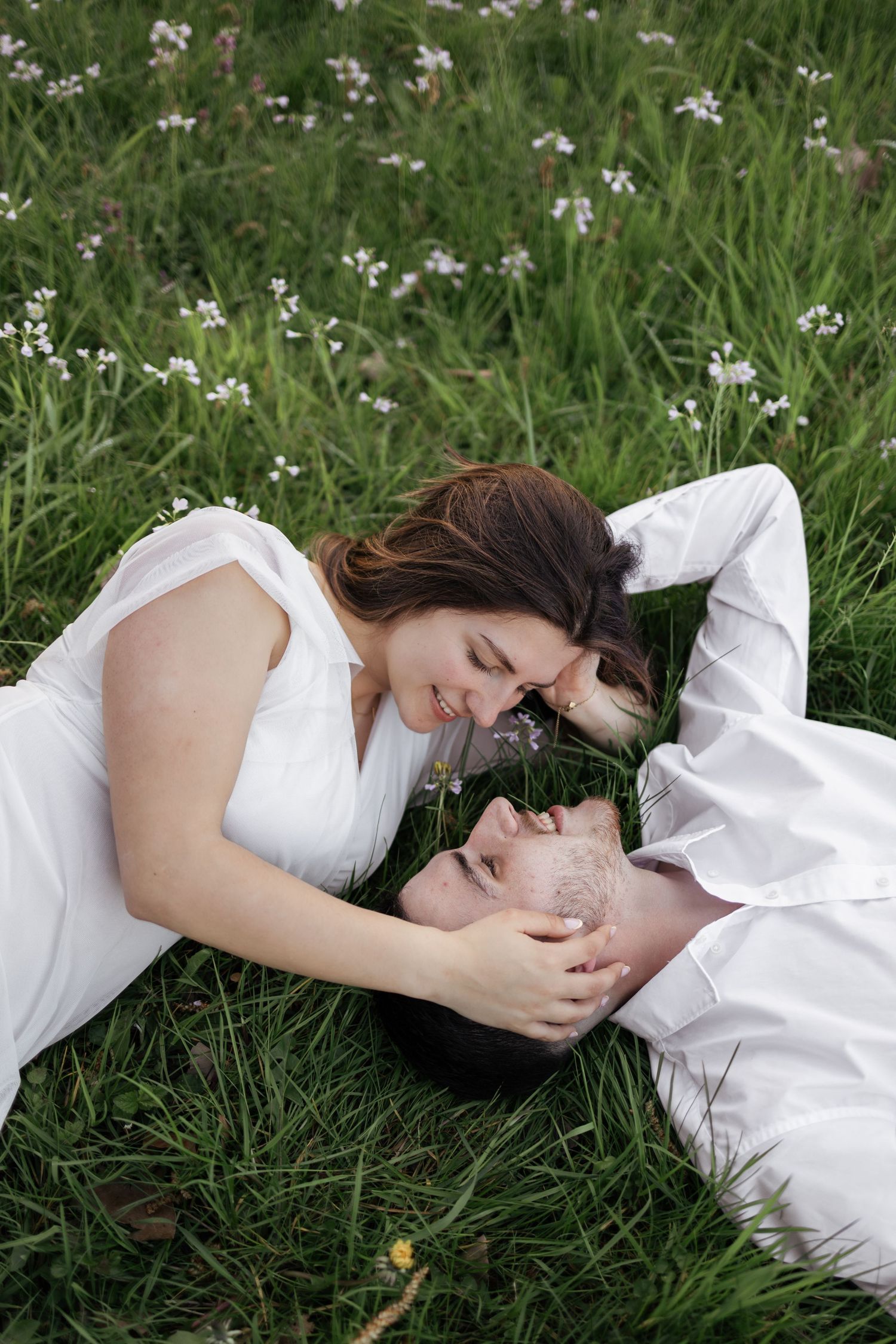 Romantic couple in white clothing lying in green grass field with wildflowers, smiling and gazing at each other.
