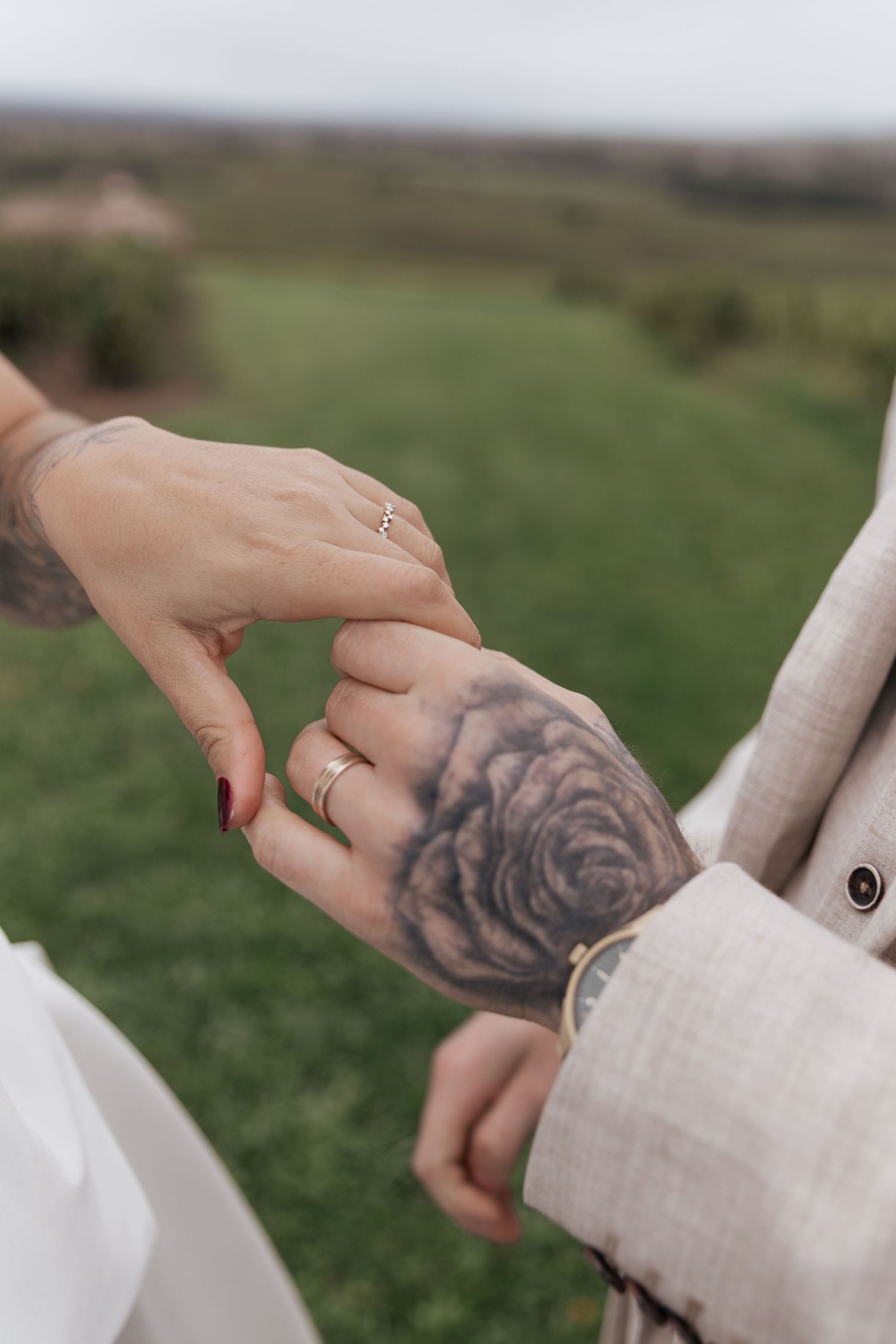 Two hands with wedding rings interlock fingers, one adorned with a rose tattoo, against a lush green landscape backdrop.