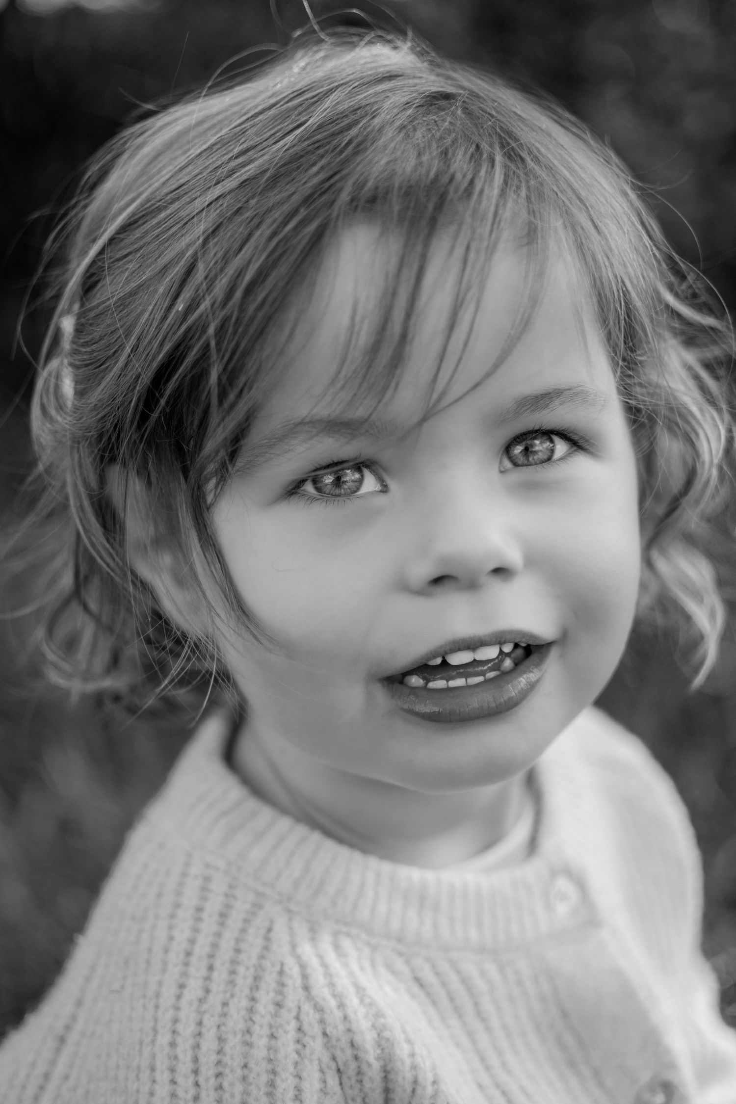 Black and white portrait of a smiling toddler girl with curly hair wearing a knit sweater outdoors.