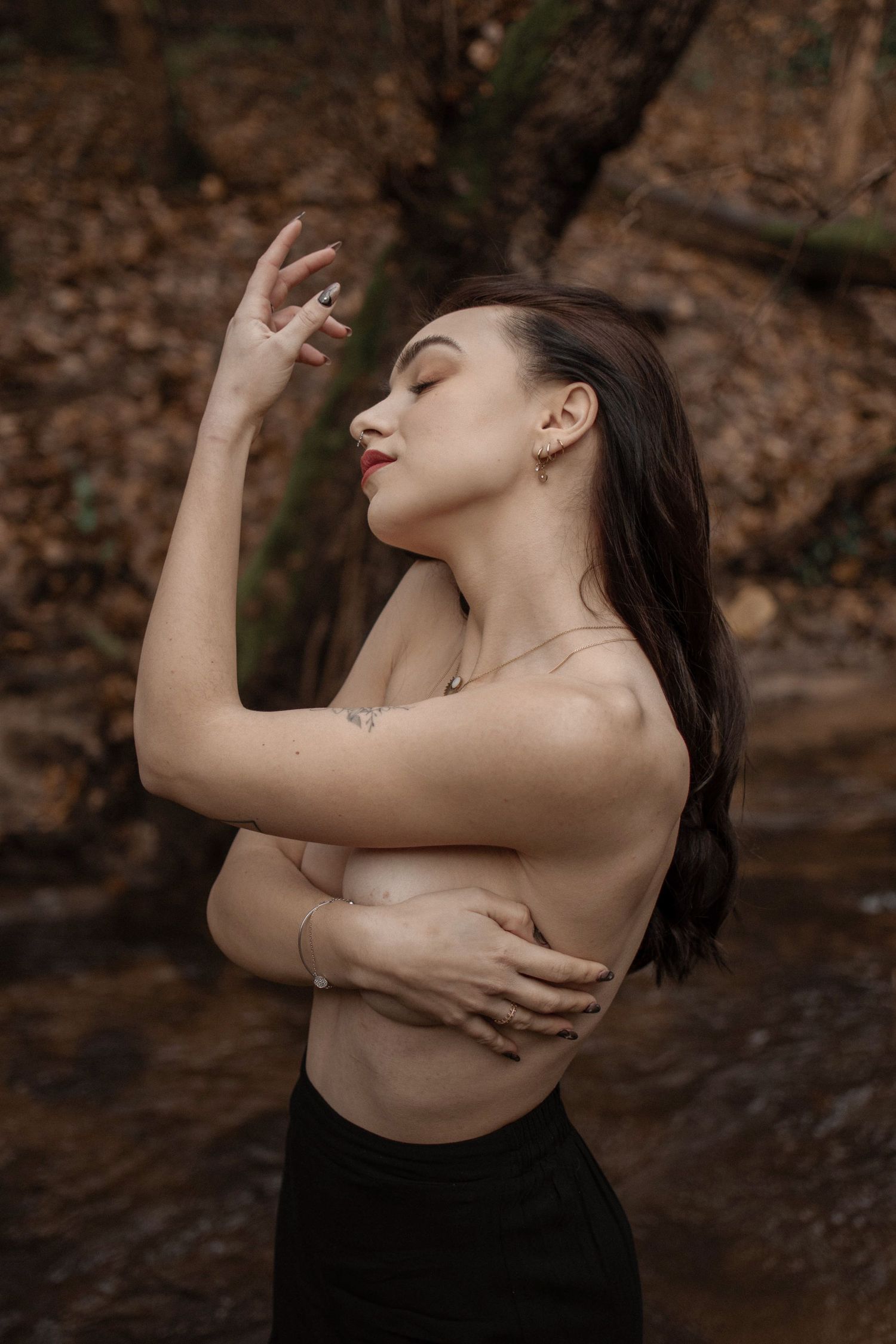 A woman with dark hair poses topless outdoors, arms crossed, eyes closed, wearing jewelry, with autumn foliage in background.