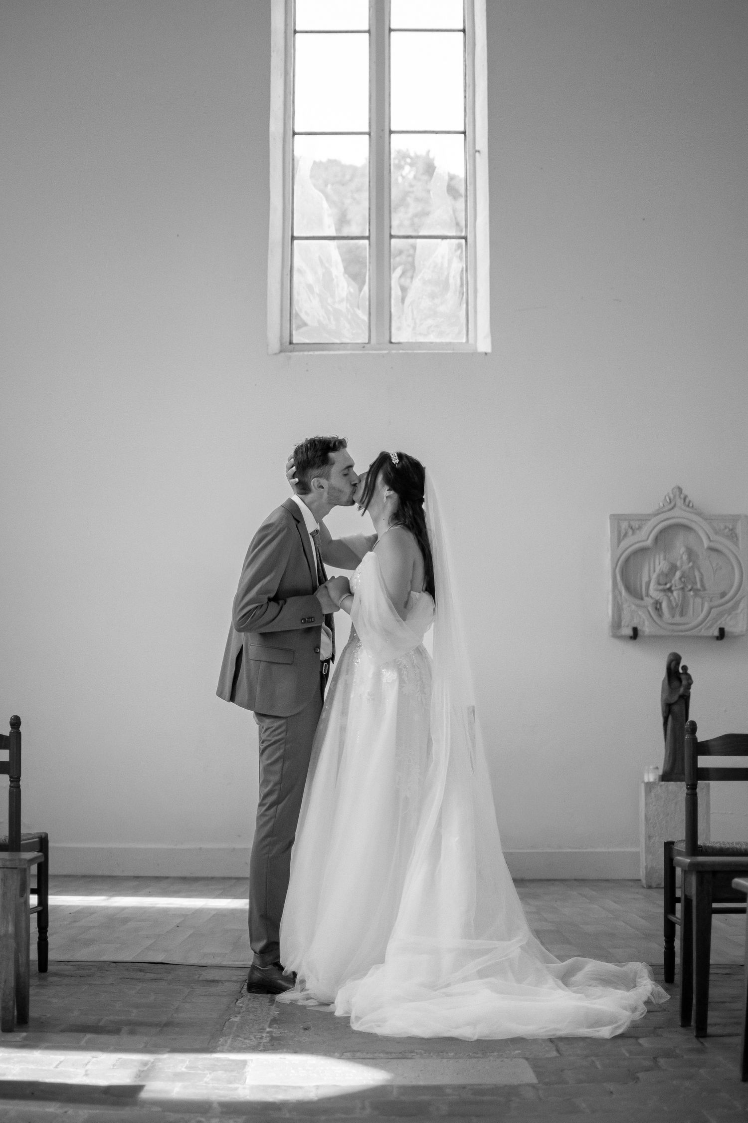Black and white photo of a bride and groom sharing a kiss inside a sunlit church, with pews and a decorative relief on the wall.