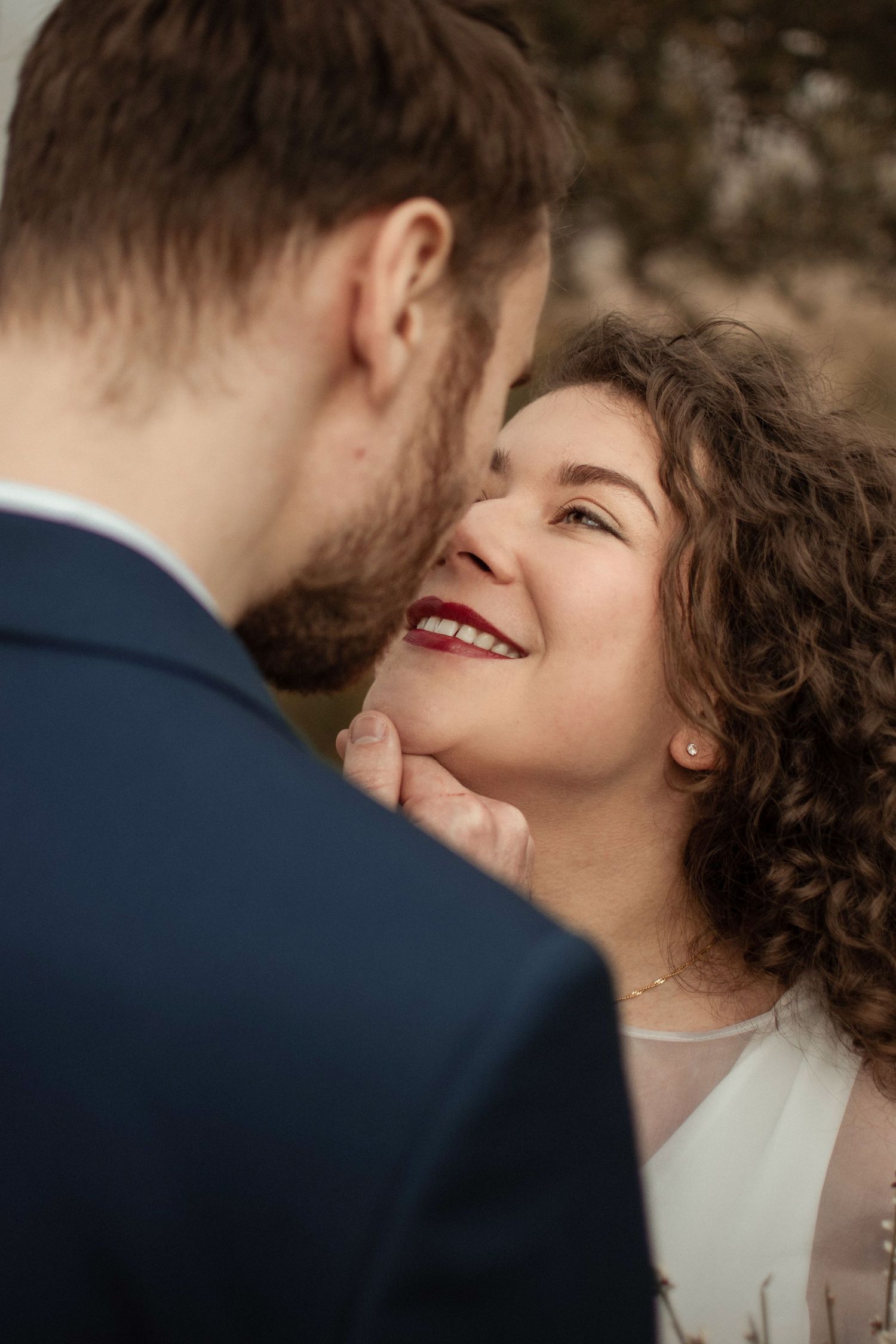 A smiling bride with curly hair and red lips gazes lovingly at a groom in a navy suit during an outdoor wedding.