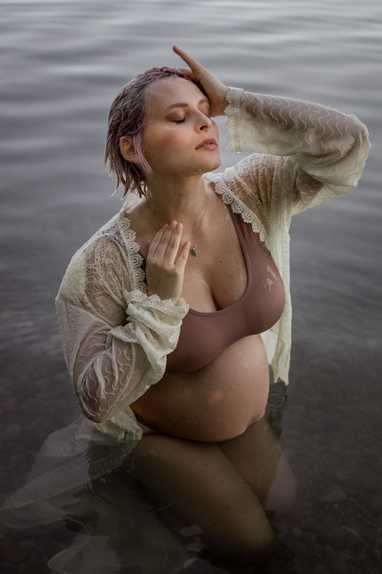 Pregnant woman in lace robe and bikini posing gracefully in calm water with eyes closed.