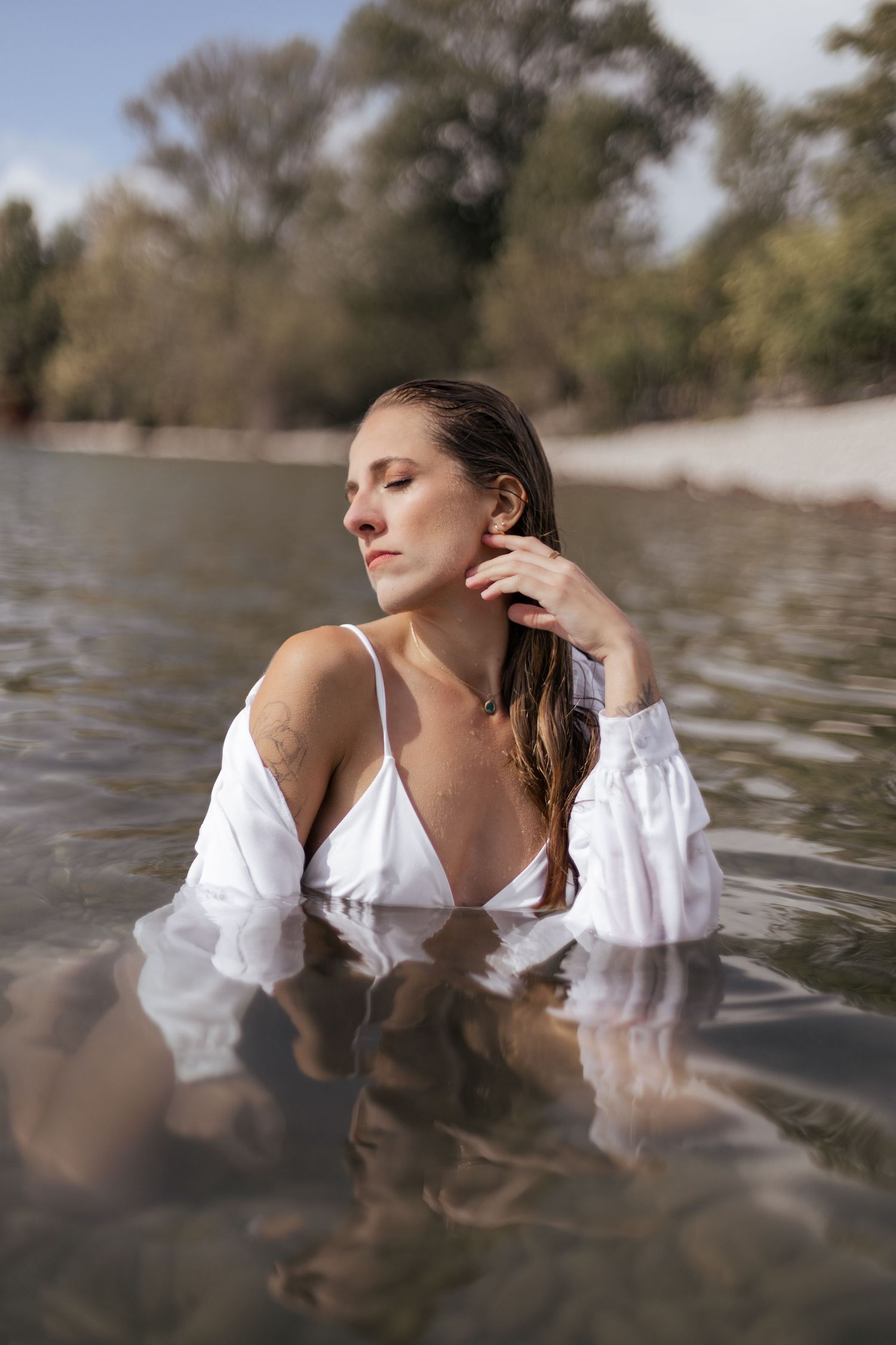 Woman in white blouse standing in calm river water with trees in background, eyes closed, hand touching neck.