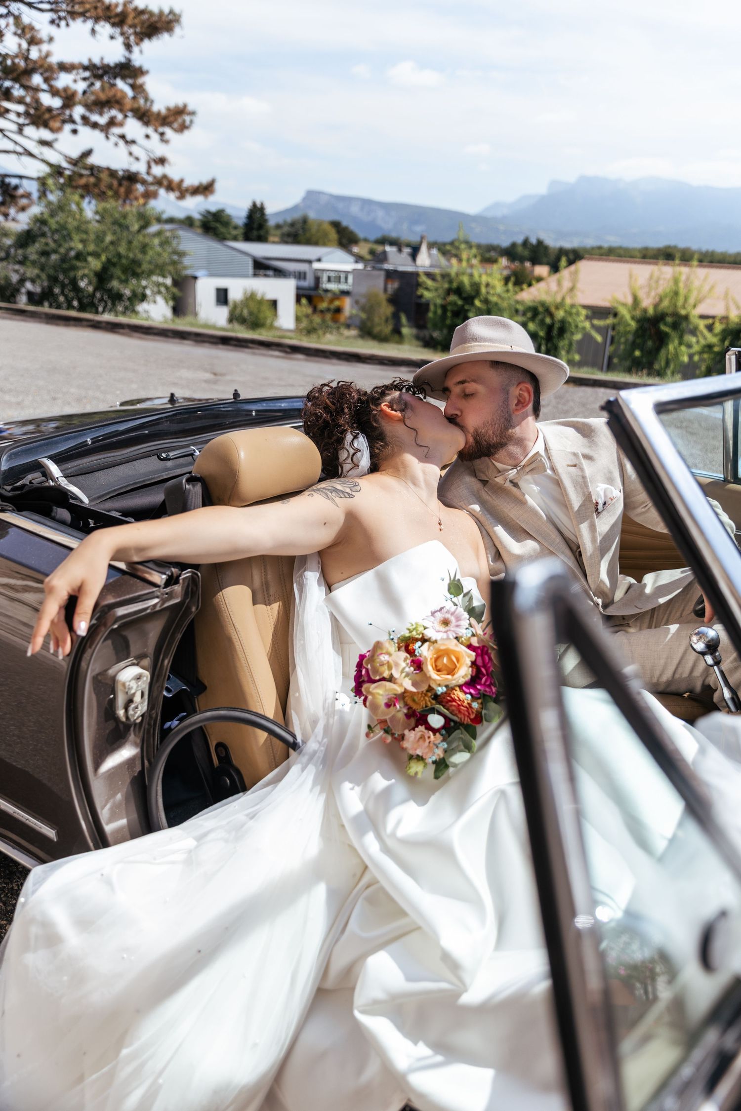 Bride and groom kissing in a vintage convertible car, bride holding colorful bouquet, mountains in background.