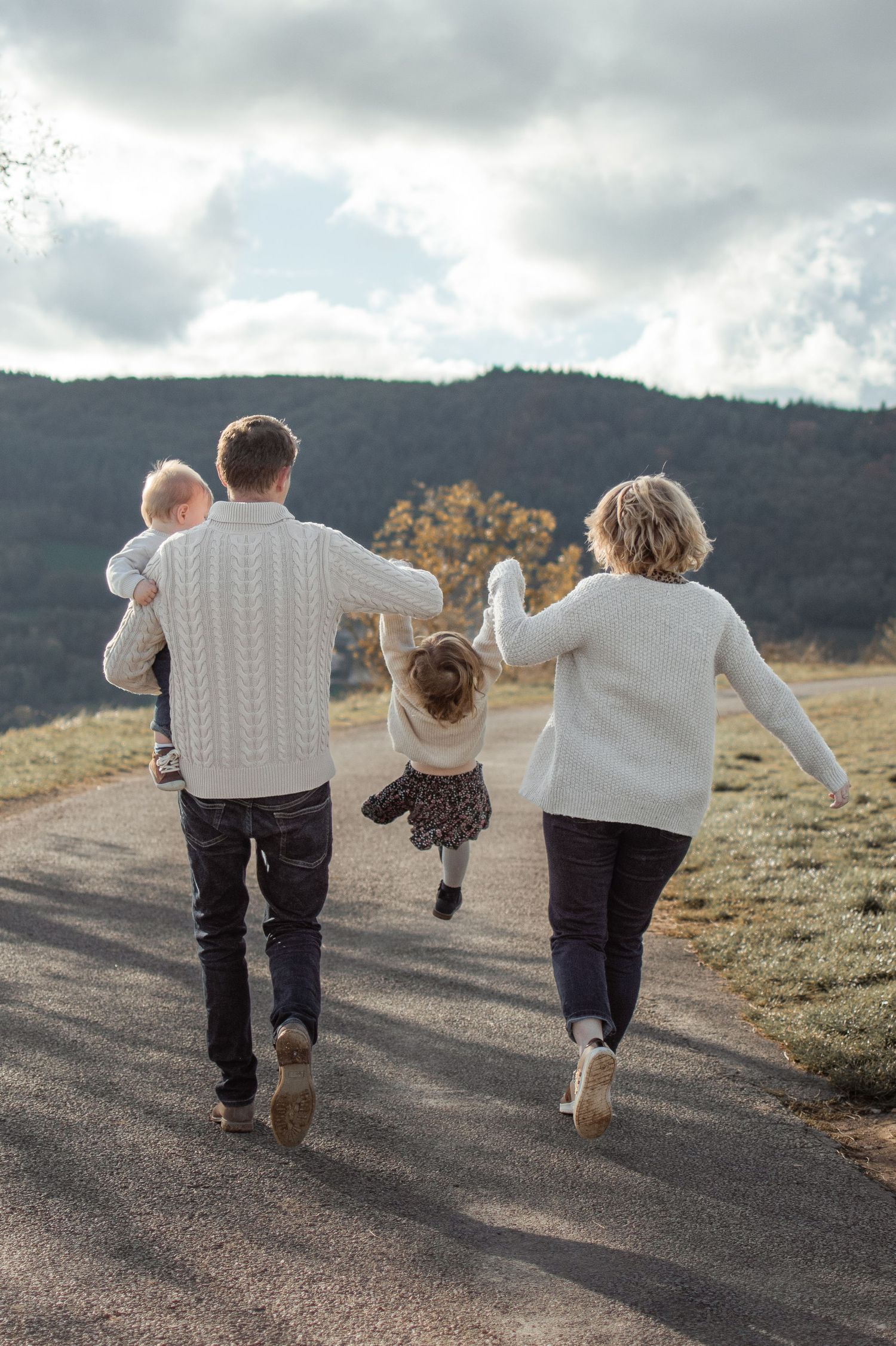 Family of four walking away on a rural road, parents swinging child between them, hills in background.