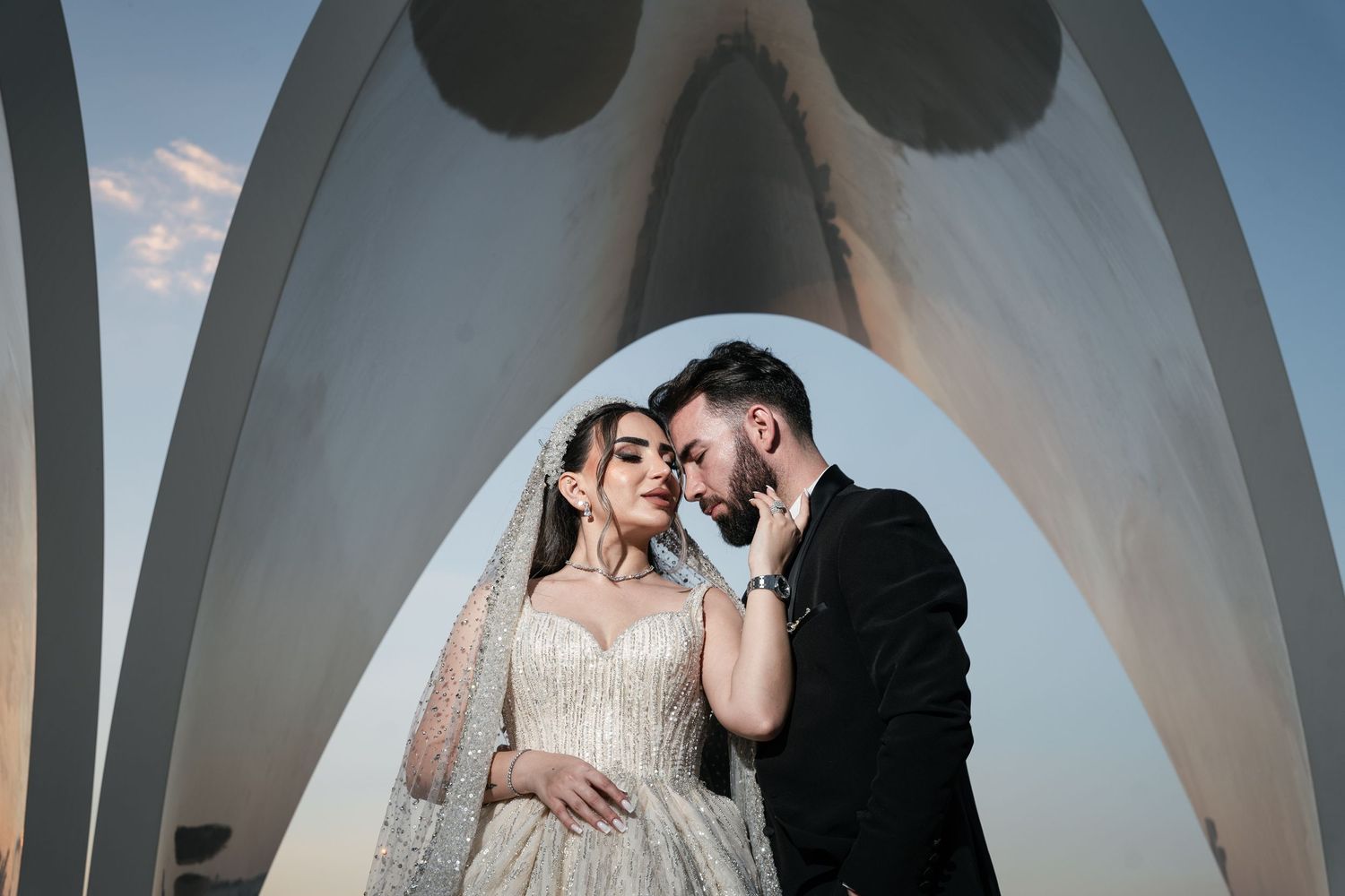 Romantic wedding portrait under a modern white architectural arch at sunset.