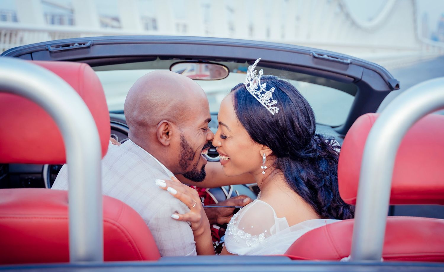 Happy couple embracing in red convertible car during wedding photoshoot.