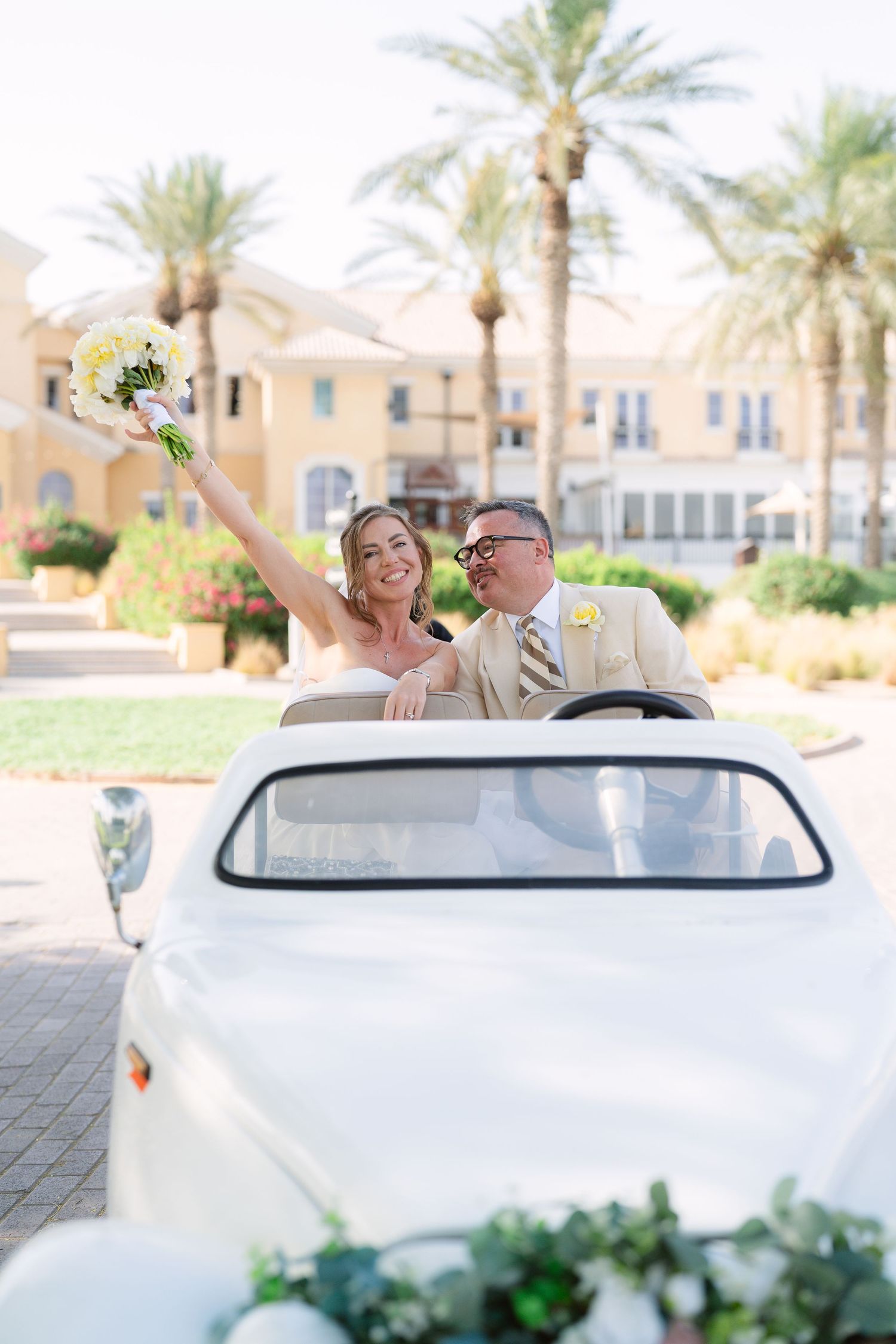 A newlywed couple celebrates in a white vintage car decorated with flowers, with palm trees and buildings in the background.
