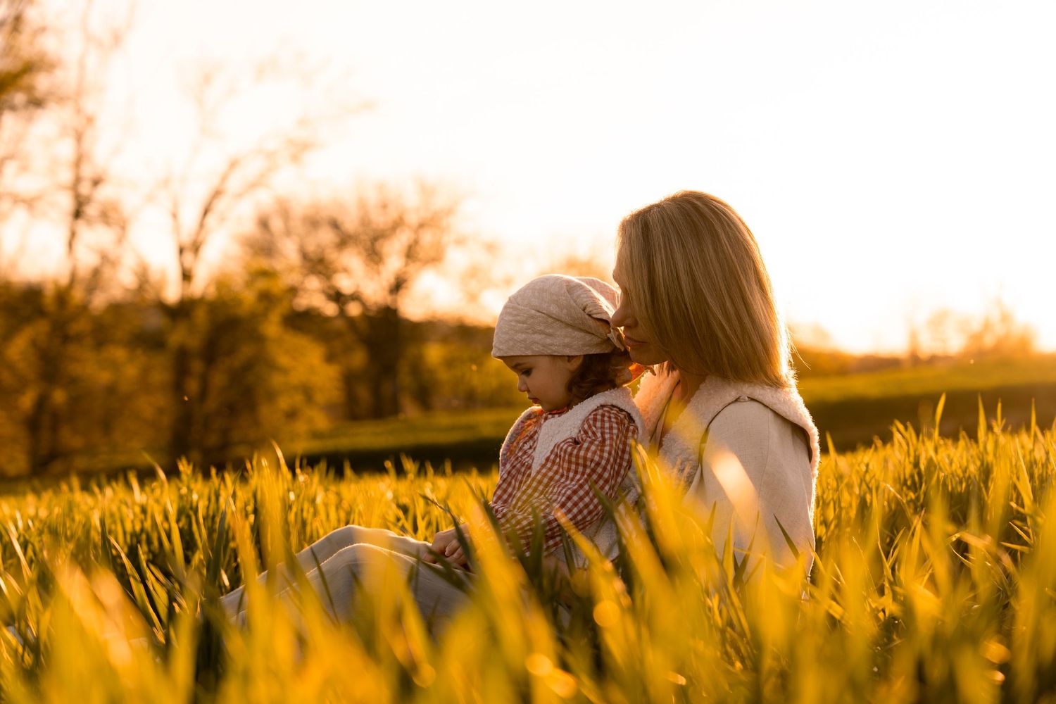 Mother and child sharing a tender embrace in a golden sunlit field at sunset, surrounded by tall green grass.