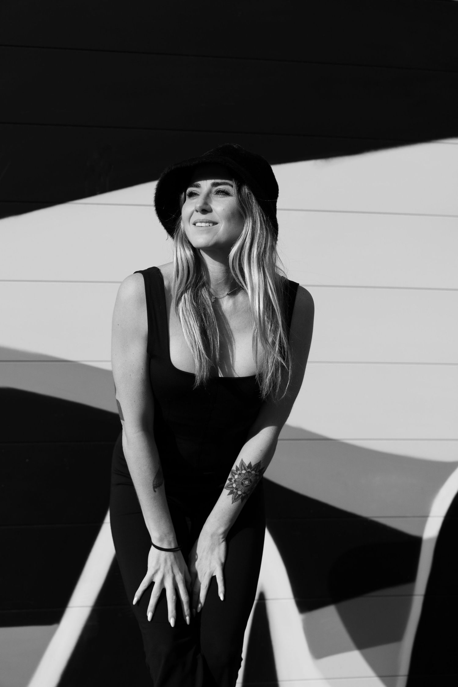 Candid black and white portrait of person in hat smiling against white wall.