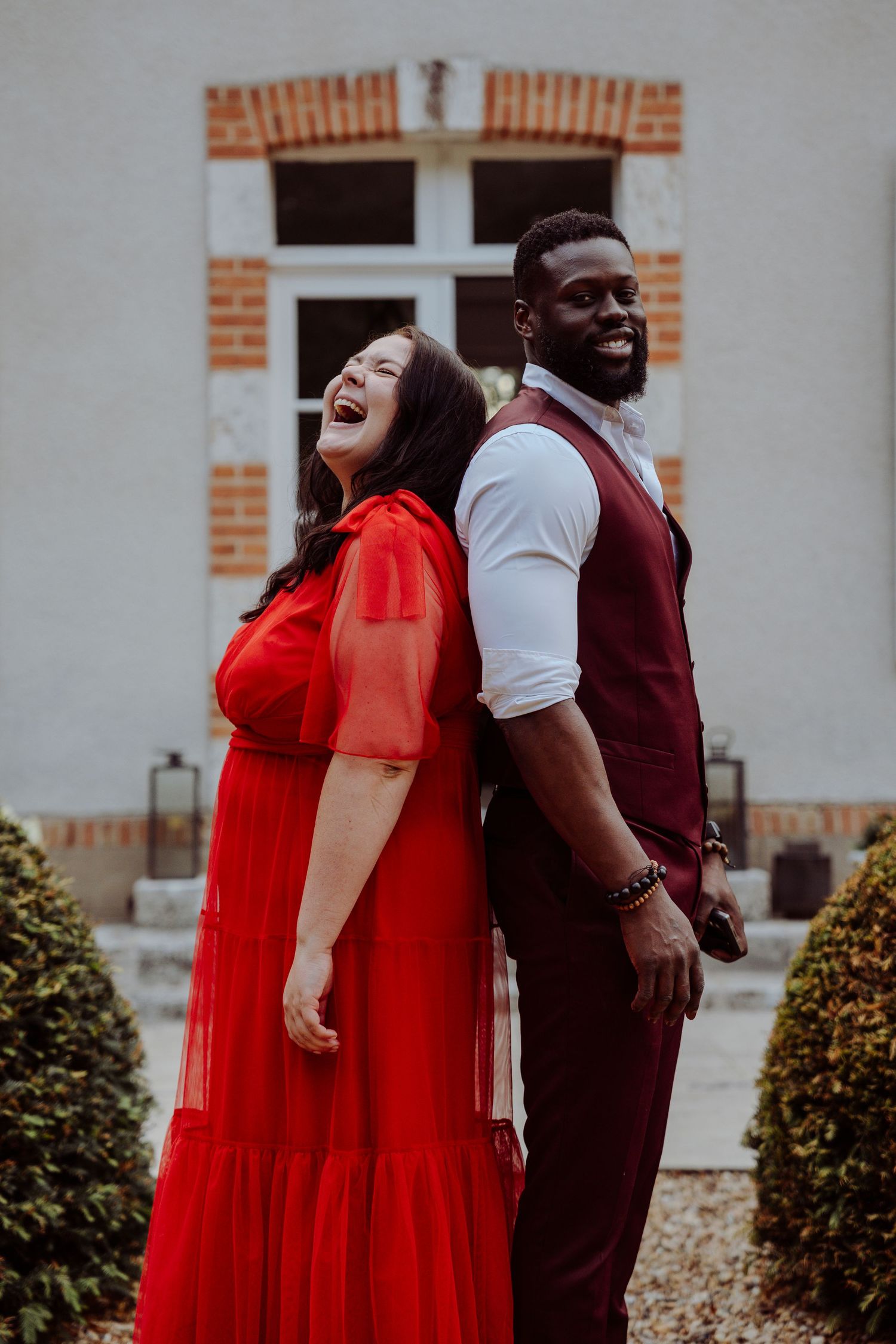 Reportage de mariage vivant et authentique. Un couple en tenue de soirée partage un moment de joie devant un bâtiment en briques.