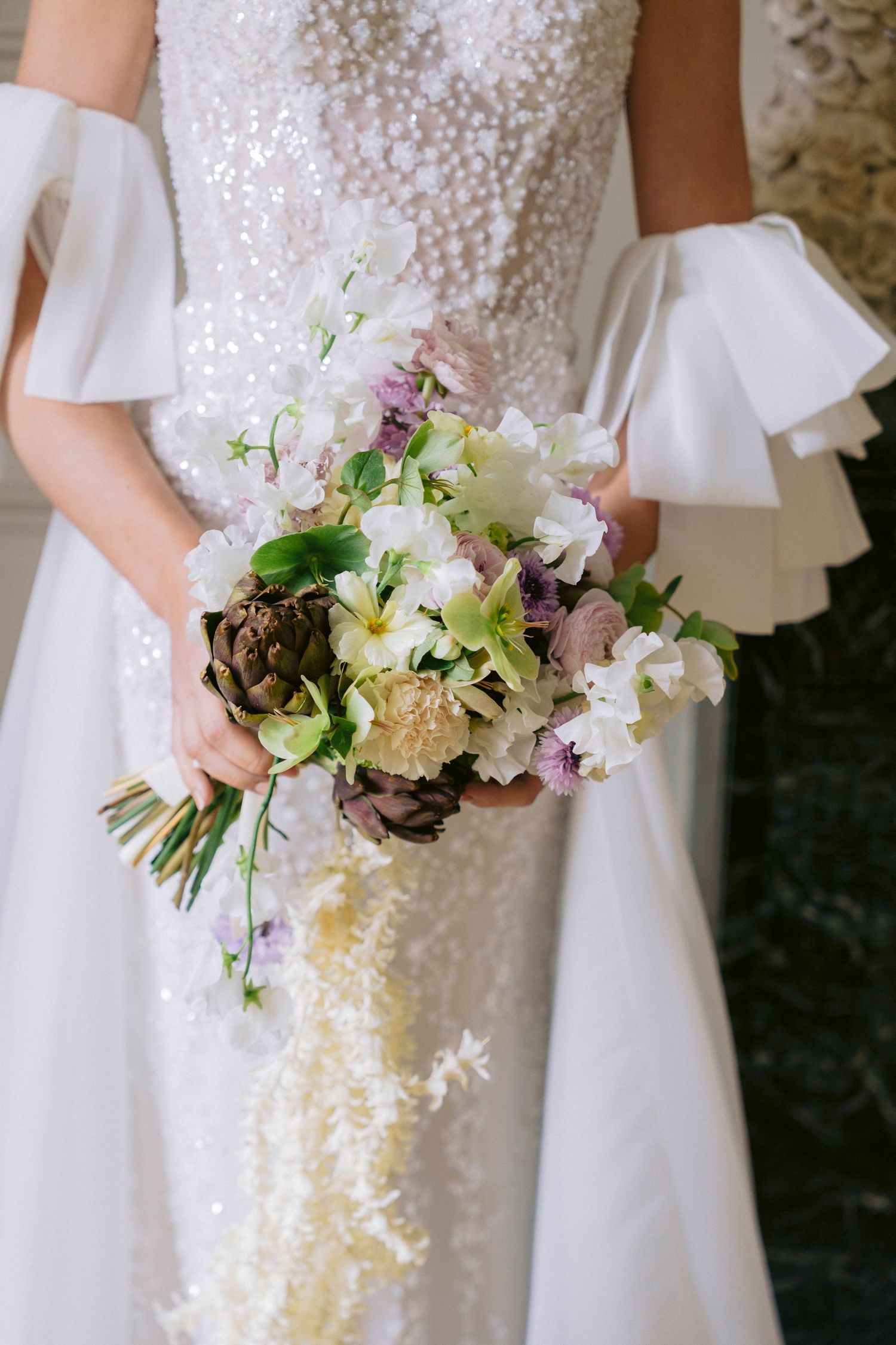 Bride in sequined white gown with ruffled sleeves holding lush bouquet of artichokes, orchids, and wildflowers.