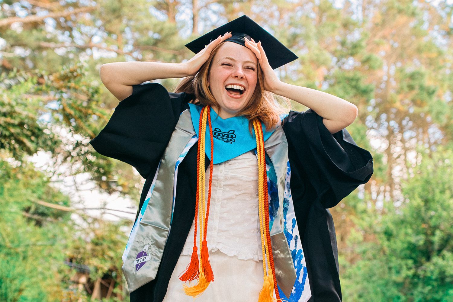 Excited young woman in black graduation cap and gown with colorful stoles laughing outdoors.