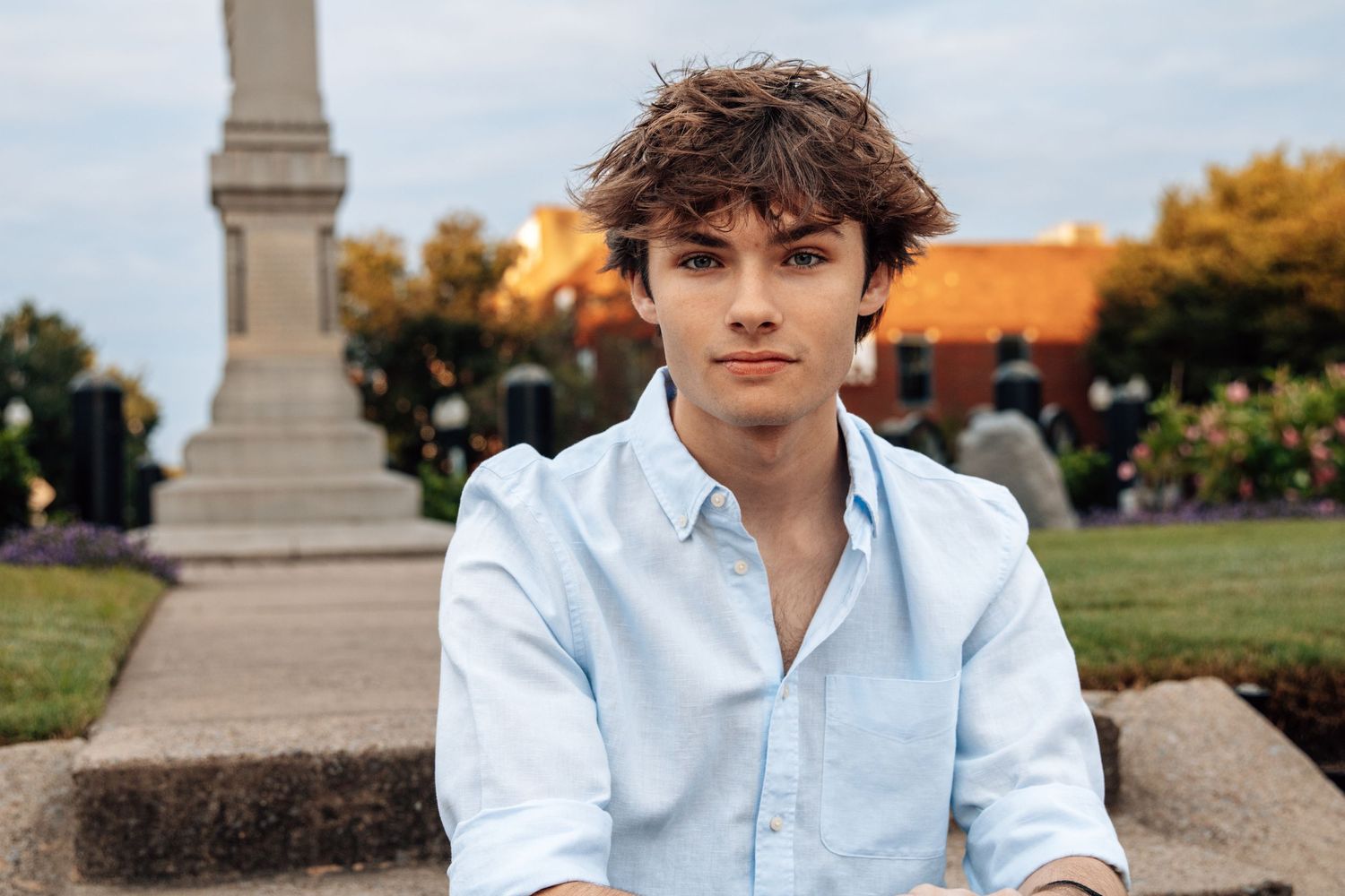 Young man with curly hair wearing a light blue shirt sitting outdoors near a monument in a park.
