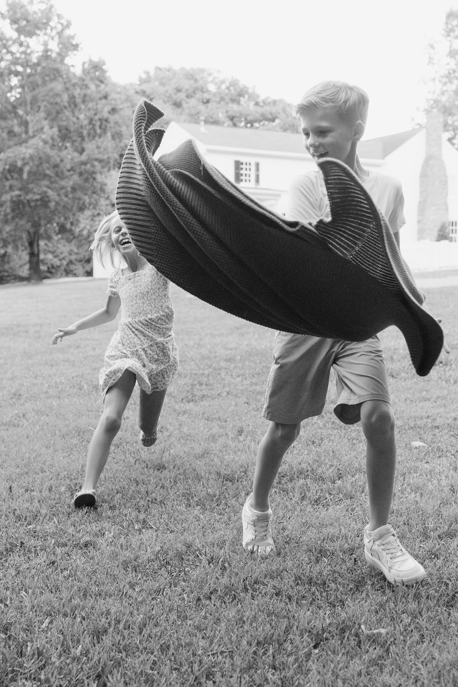Two children playing outdoors on grass, boy running with blanket cape as girl chases him near a house.