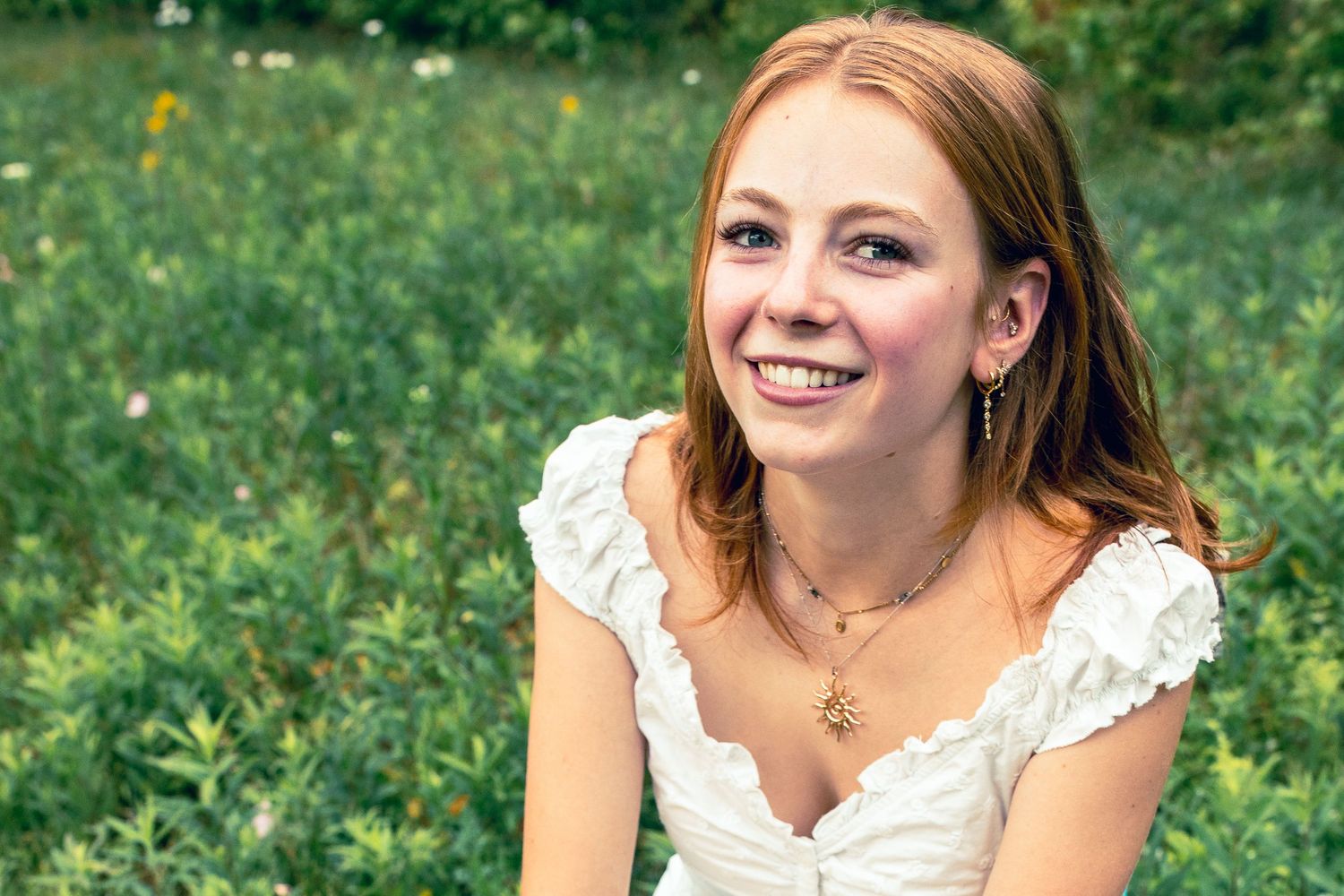 Smiling young redhead woman in white dress sitting outdoors in a green meadow with wildflowers.