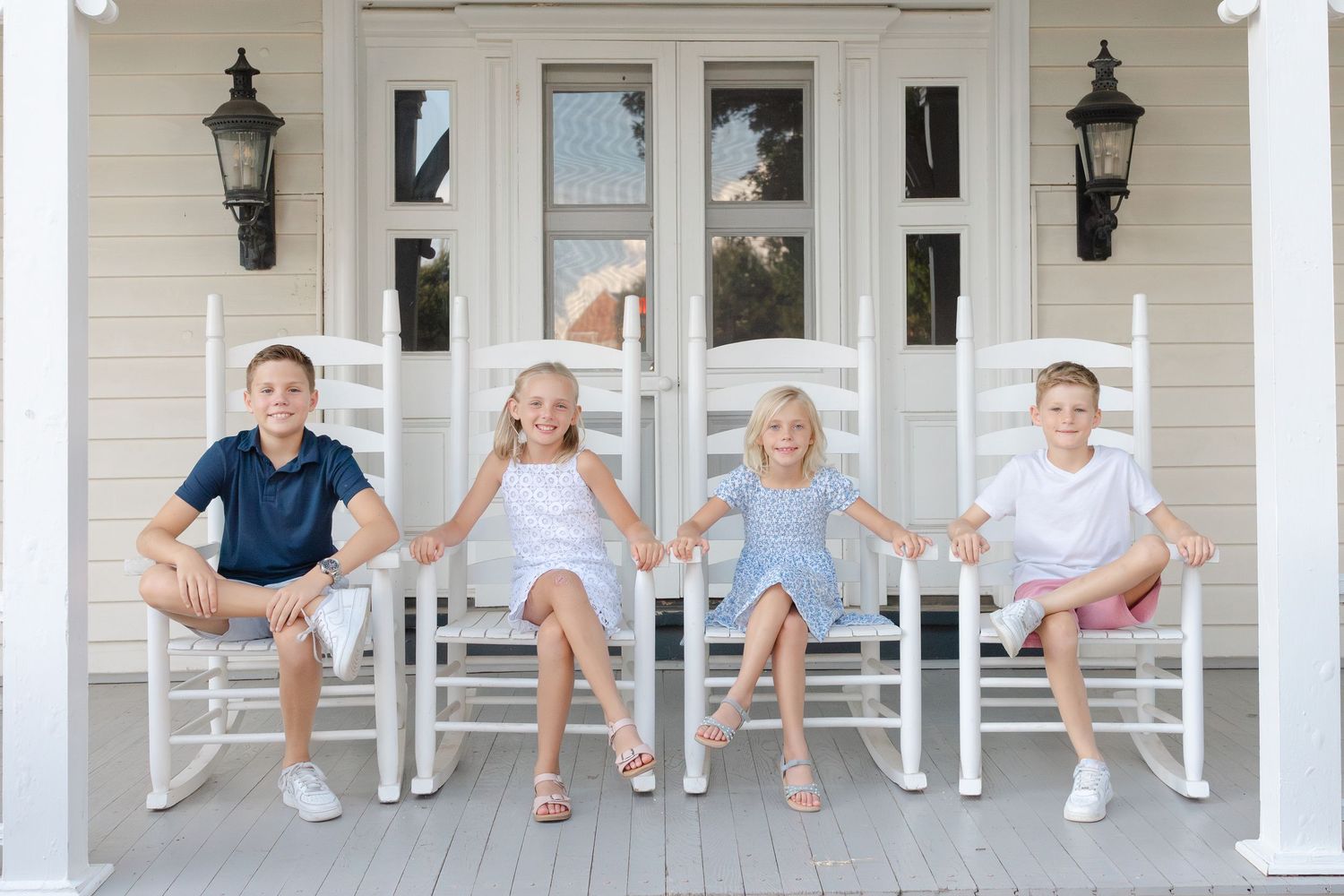 Four children sit holding hands in white rocking chairs on a classic white porch, smiling joyfully together.