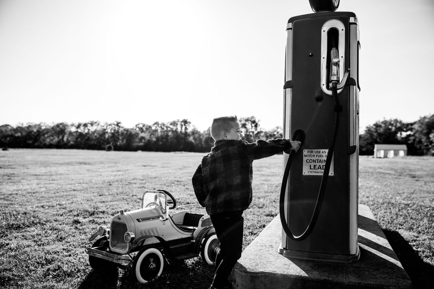 Black and white photo of a child reaching up to a vintage gas pump beside a classic pedal car in a field.