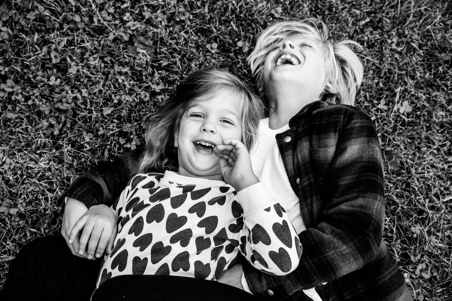 Two joyful children laughing and lying on grass in a black and white photo, wearing heart-print and plaid shirts.