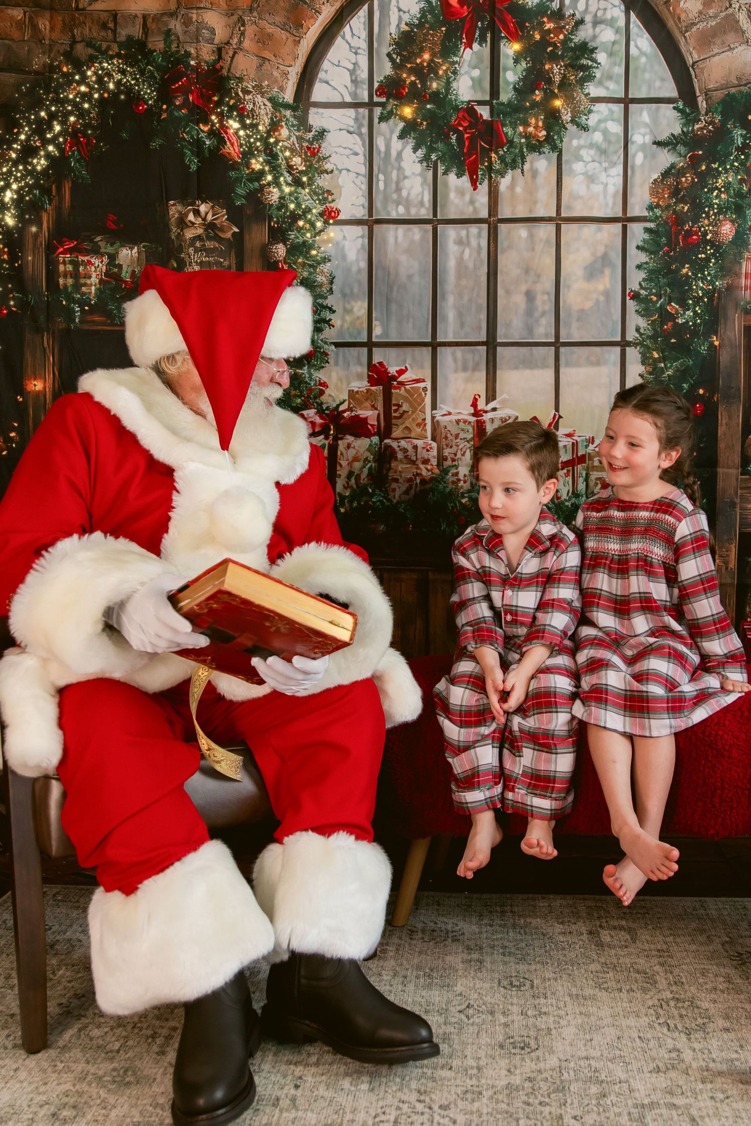 Two children in plaid pajamas sit beside Santa Claus reading a book in a festive Christmas decorated room.