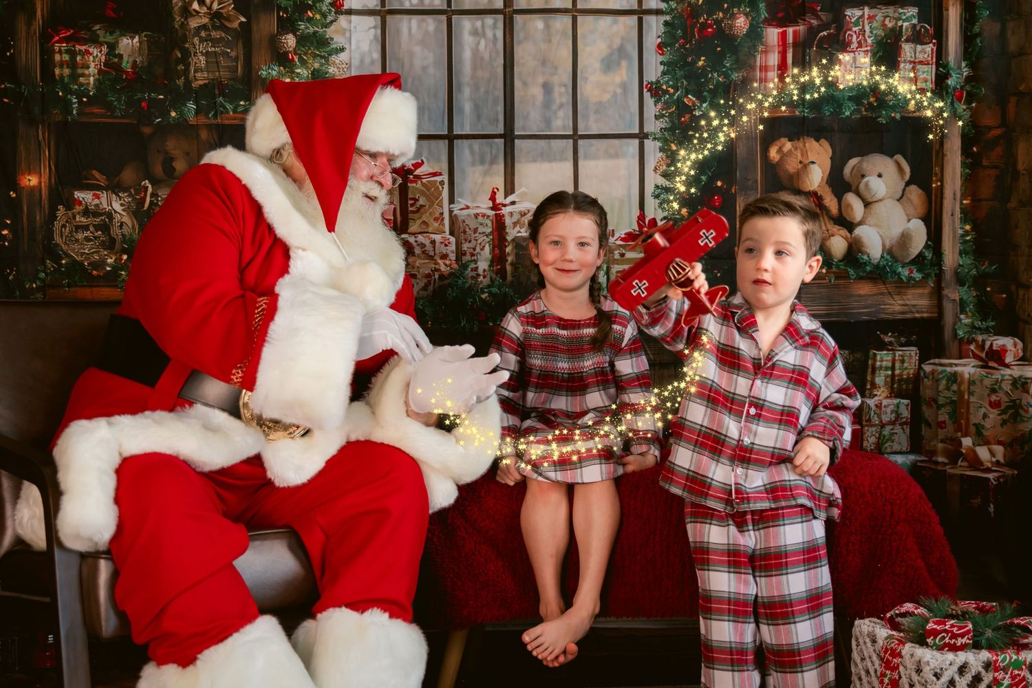 Two children in plaid pajamas pose with Santa Claus amid festive Christmas decorations and wrapped gifts.