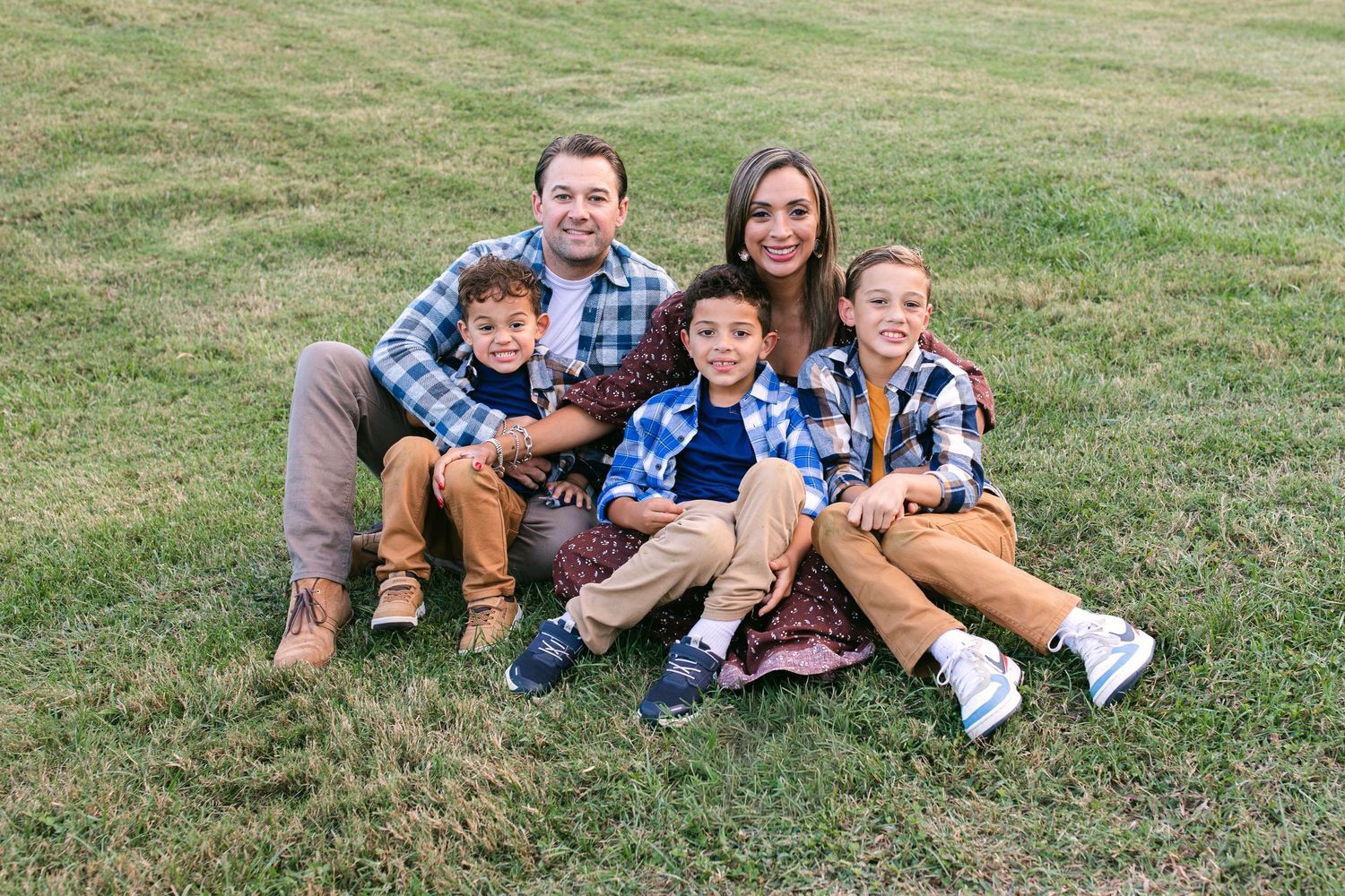 Happy family of five smiling together on green grass, parents with three young boys wearing coordinating plaid outfits.