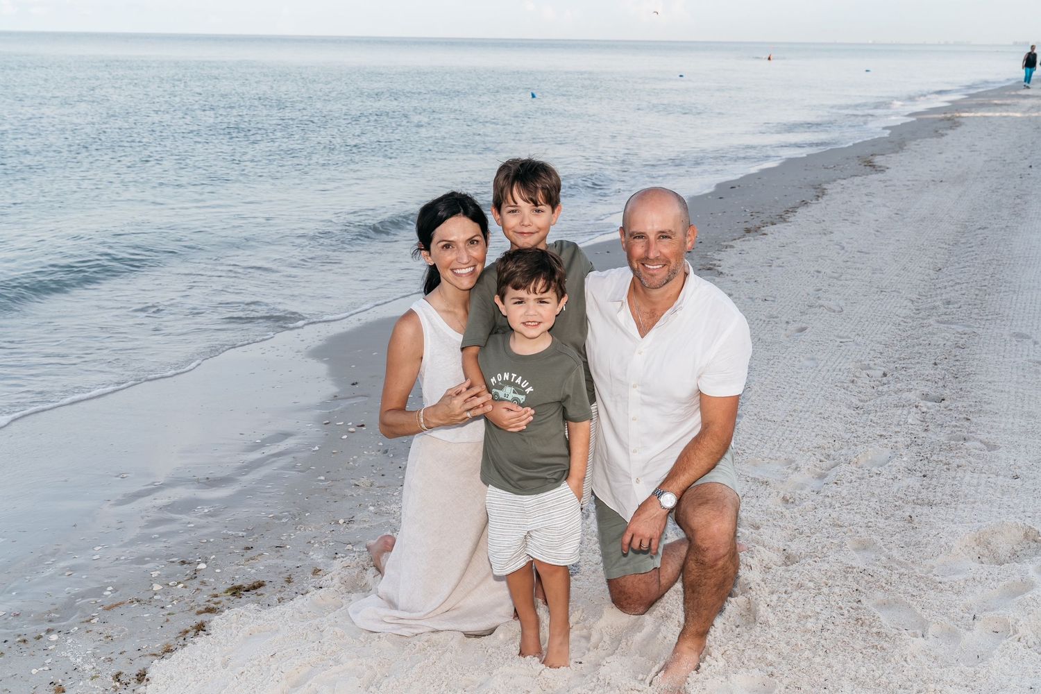 A family of four poses together on a serene beach at sunset with calm ocean waters in the background. Taken by family photographer.