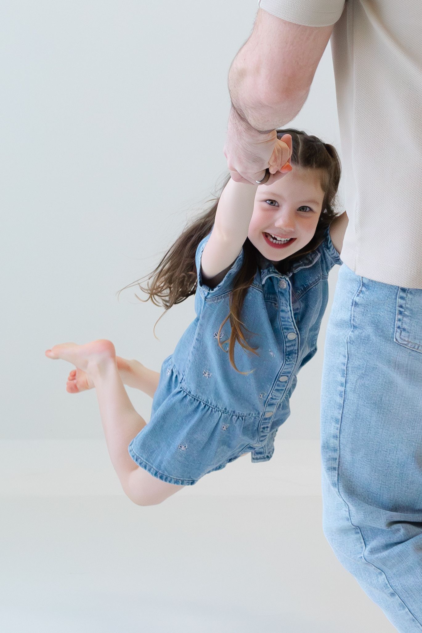 A joyful girl in a denim dress being swung by an adult's hand, laughing with hair flying in the air.