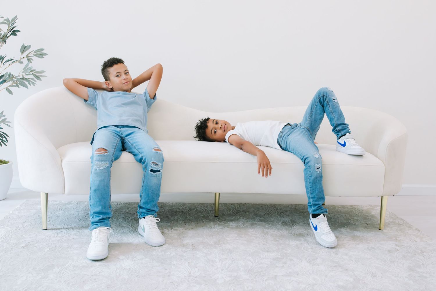 Two boys in jeans and white sneakers relaxing on a white velvet sofa in a bright modern living room.