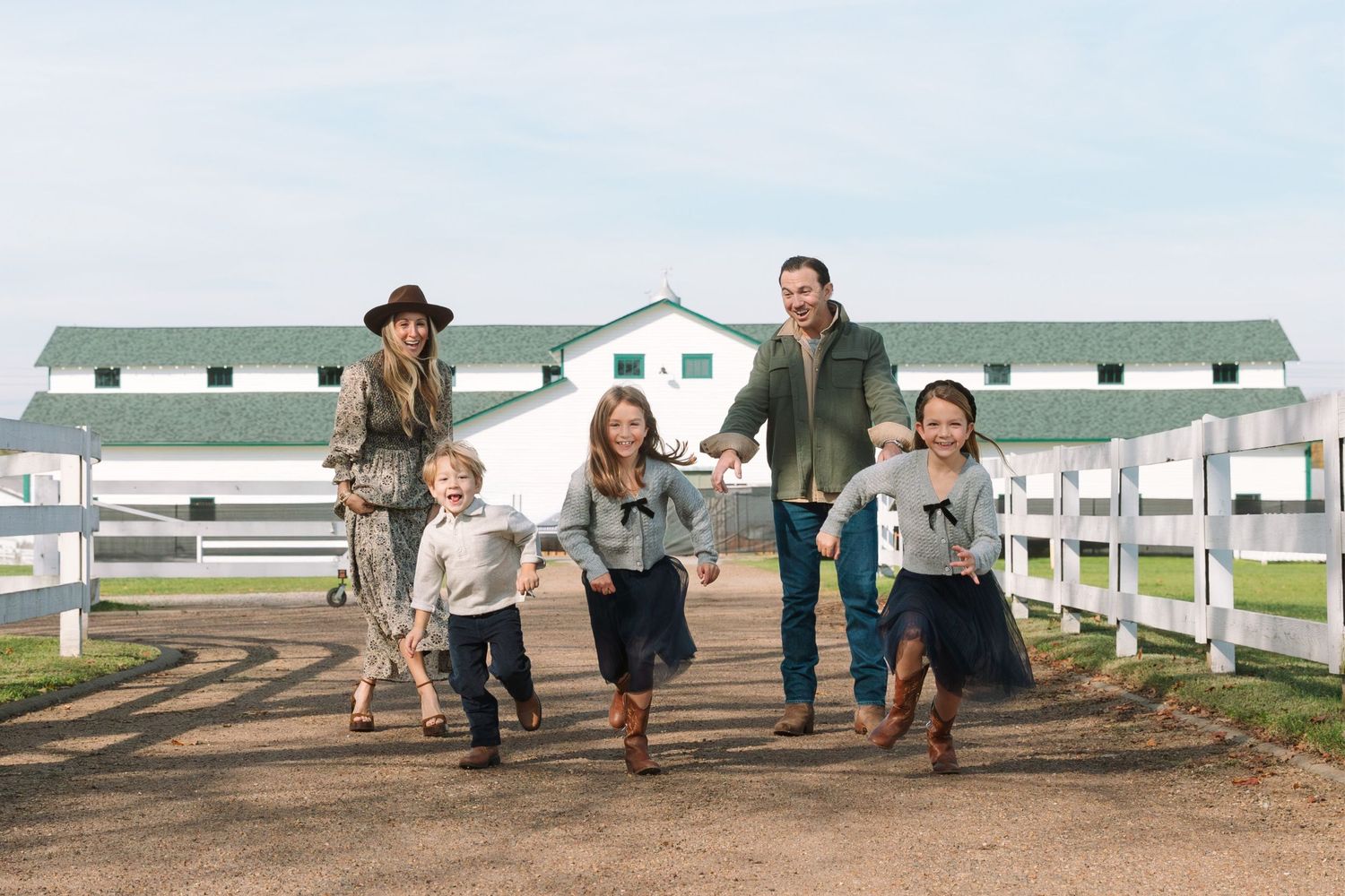 Happy family with two adults and three children walking together  with white fenced barn in background. Peachtree city family photographer.